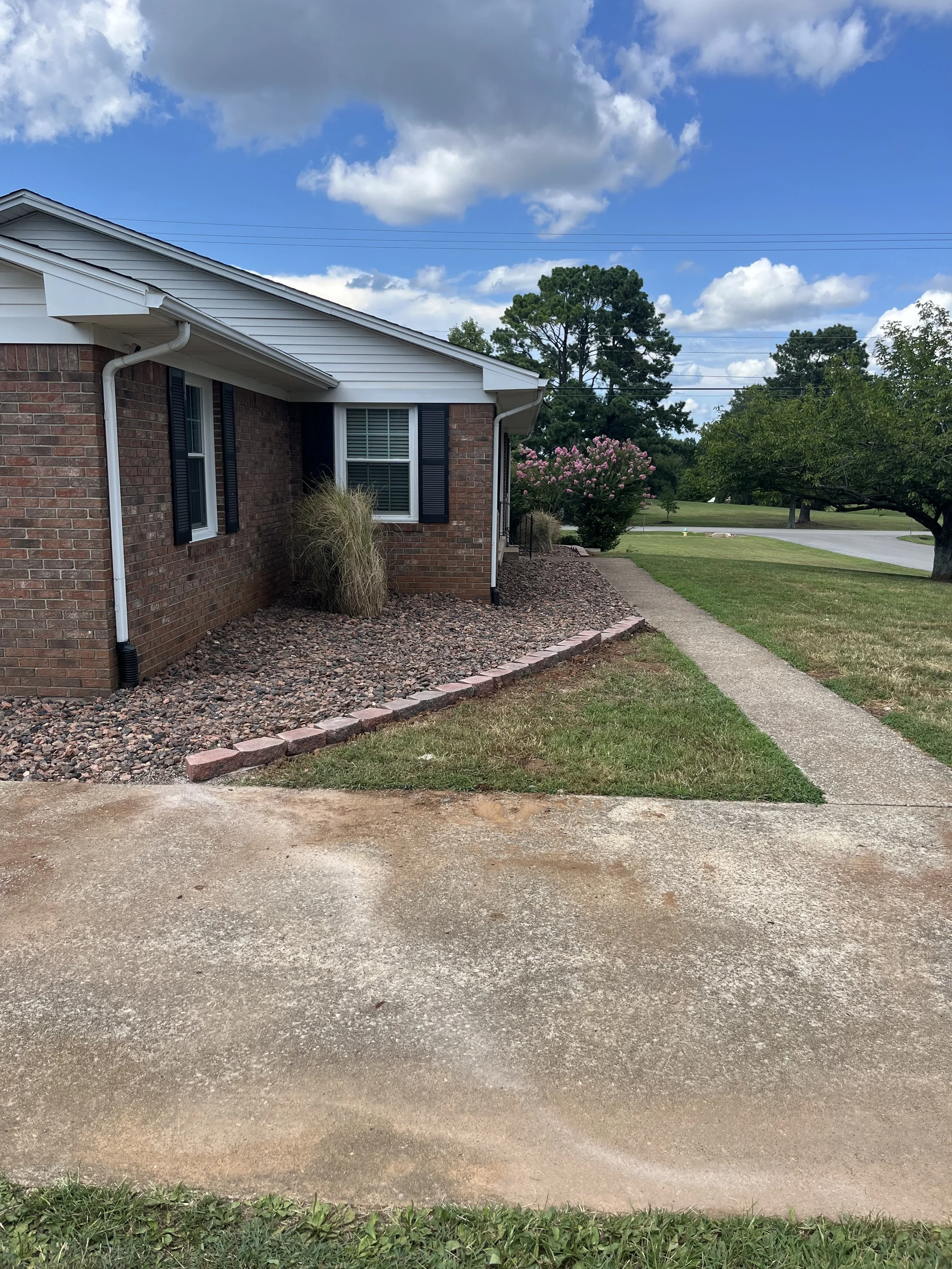 A suburban house with a brick exterior, white roof, and black shutters, surrounded by a lawn, a sidewalk, and trees under a partly cloudy sky.