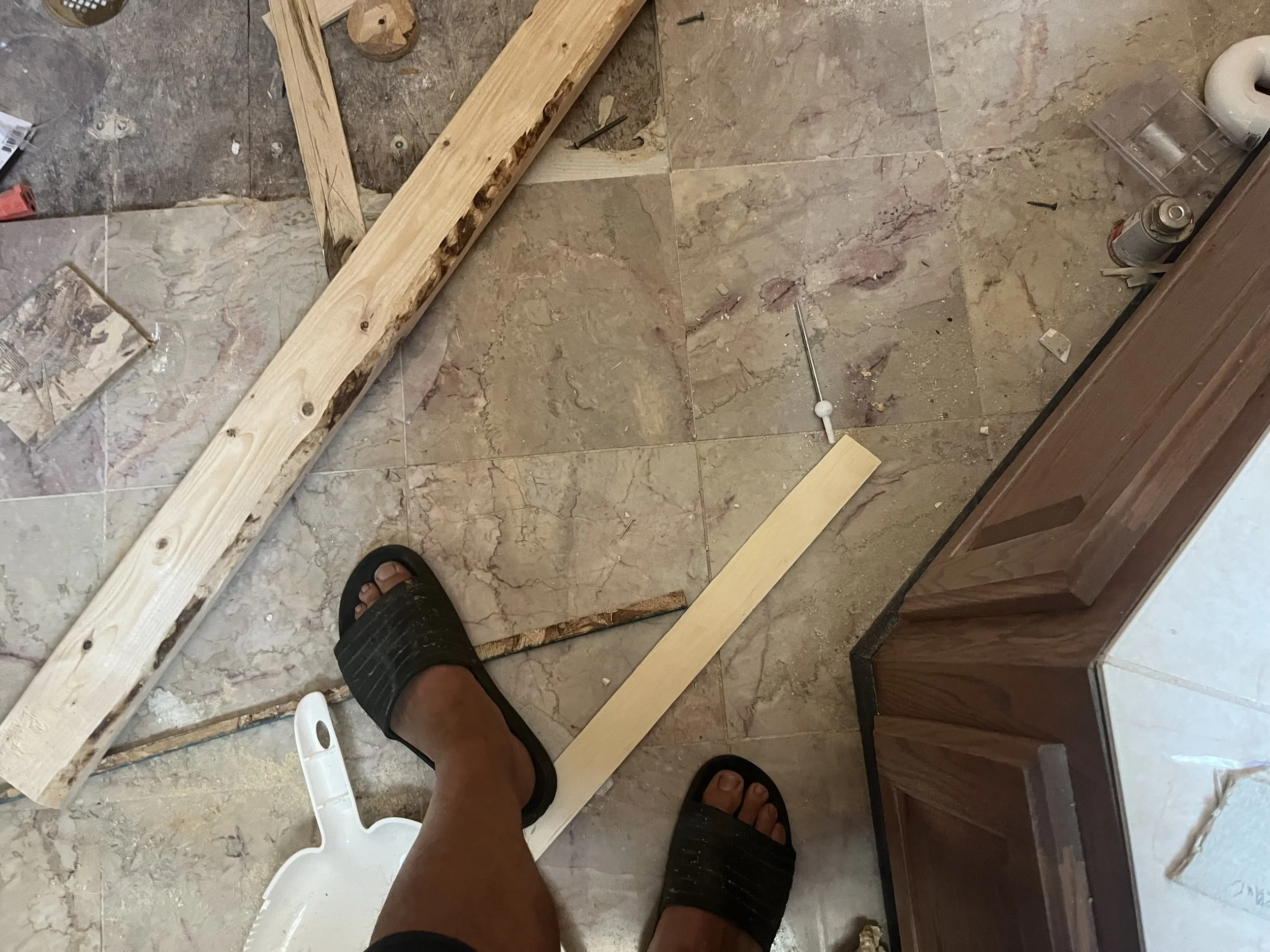 Person standing in a room with a tiled floor, wearing black slides, with partial construction work in progress. There are wooden beams, a white plastering float, and some spray cans and other construction supplies scattered around.