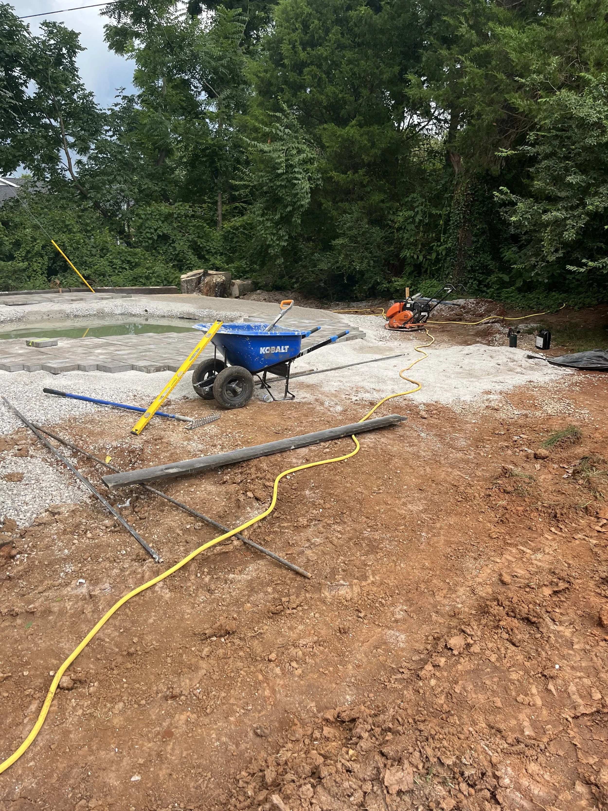 Construction site with equipment and tools near a small pond, surrounded by trees and dirt ground.