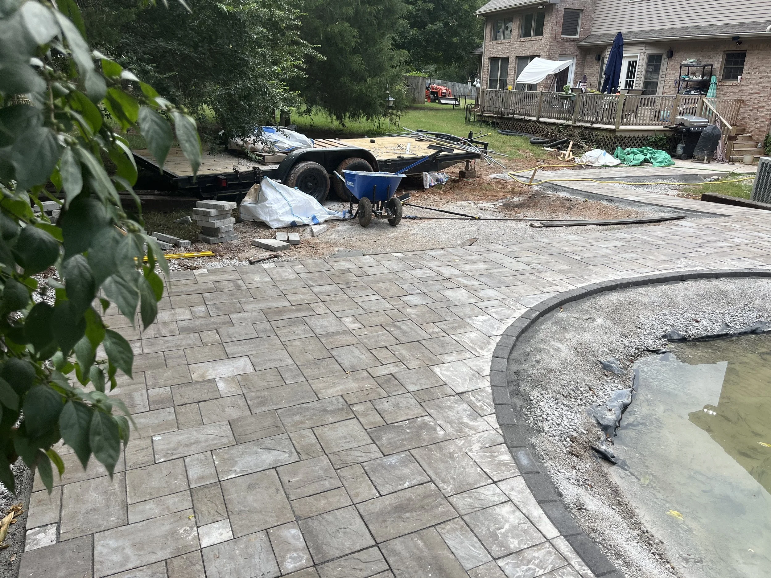 Backyard patio under construction with newly laid paver stones, construction tools, and materials scattered around, with a house and trees in the background.