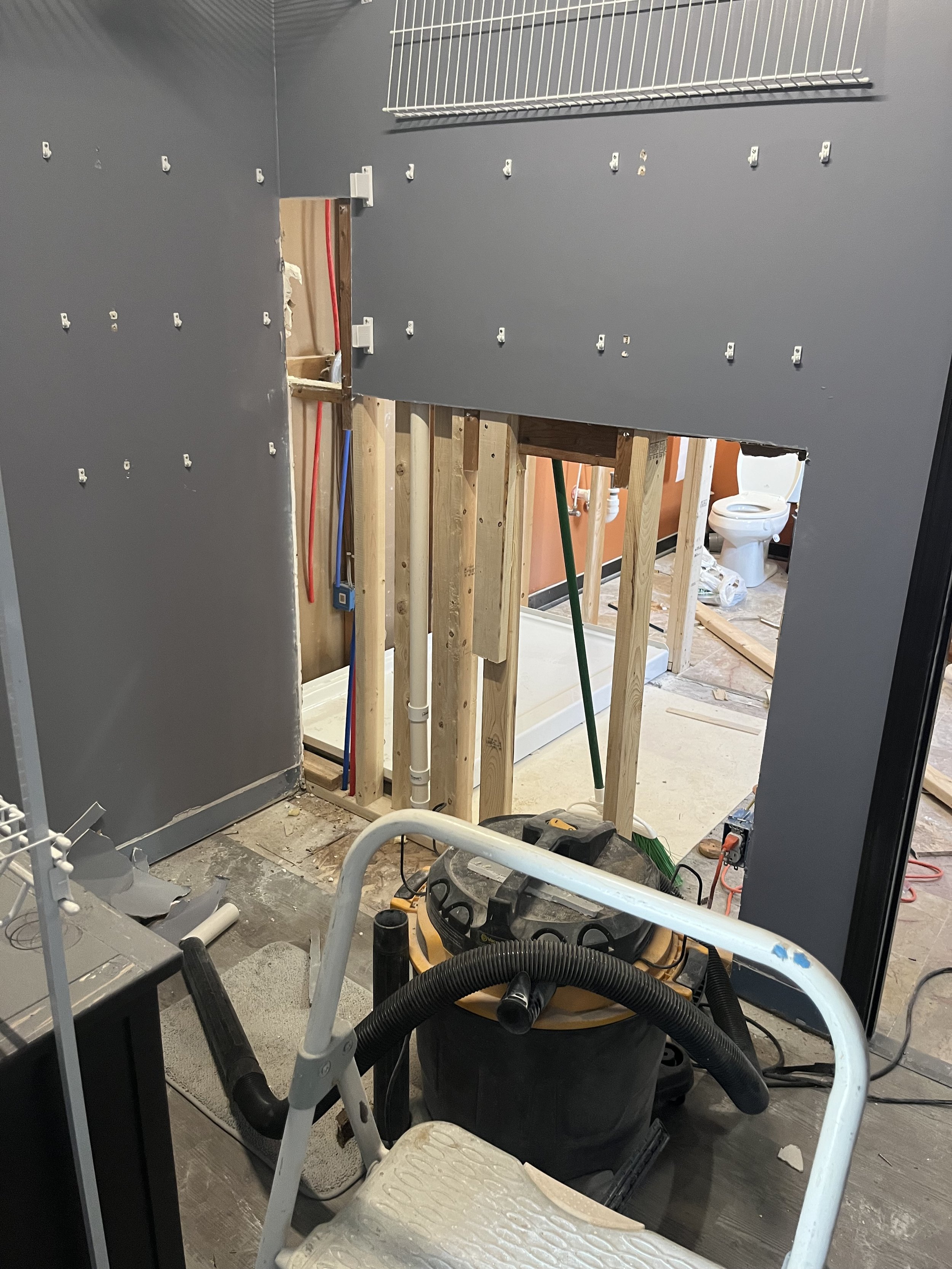 Interior of a bathroom under renovation, with exposed wooden framing and plumbing, a toilet in the background, and construction tools including a wet/dry vacuum and a ladder.