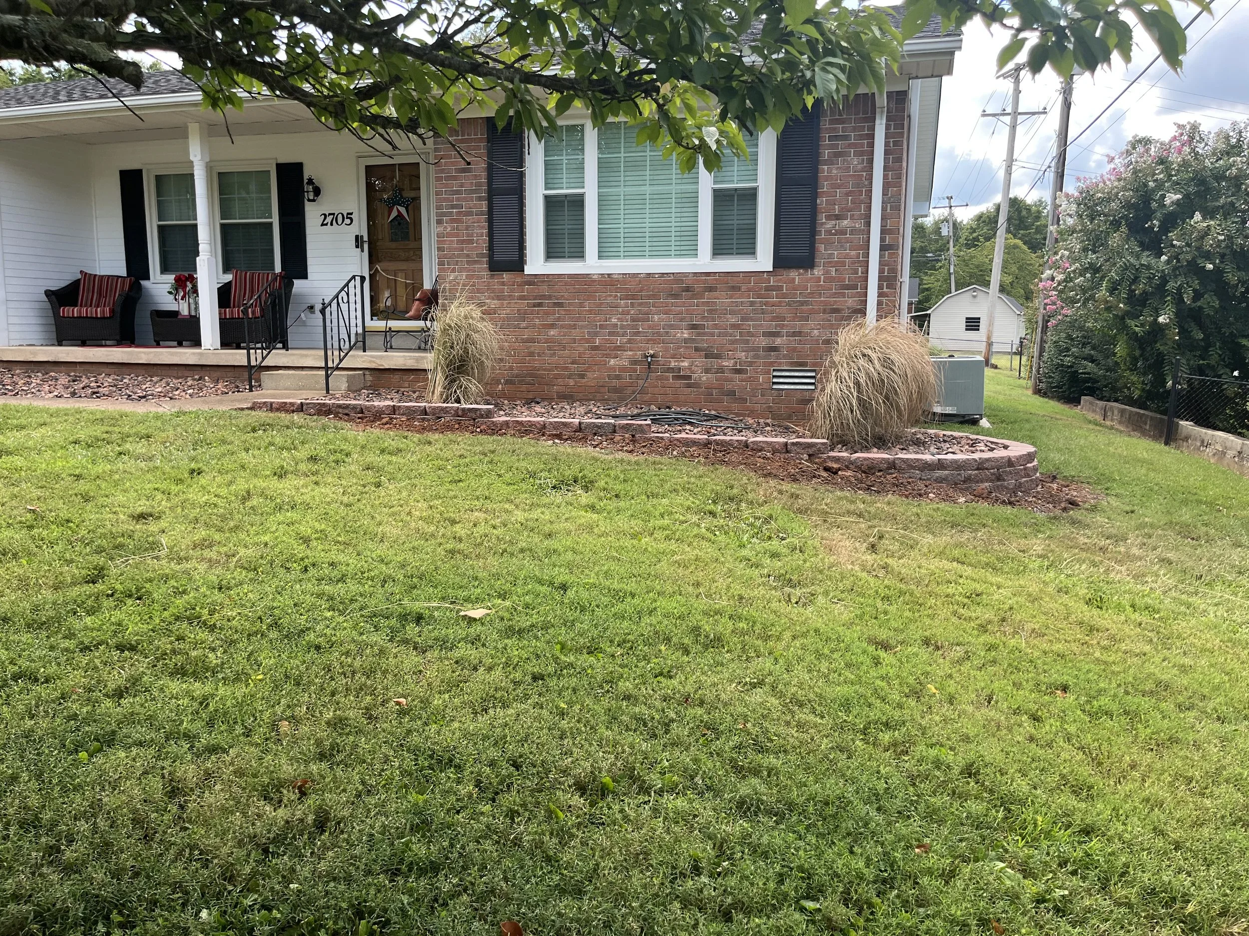 Front yard with grass and brick landscaping, house with porch, two chairs, and potted plants.