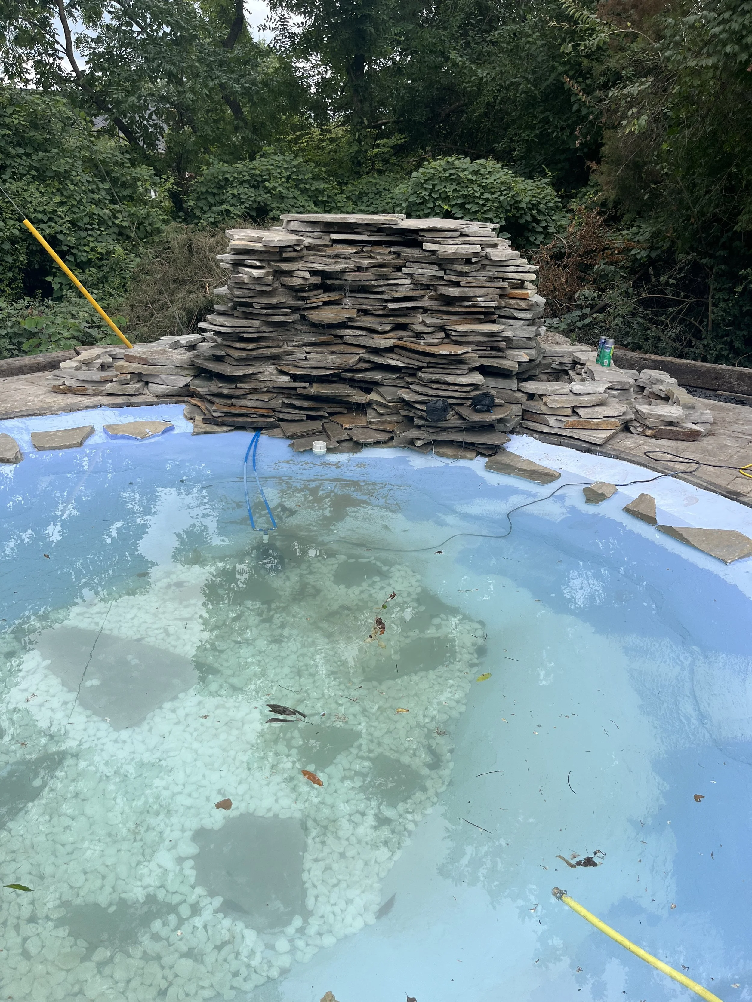 An outdoor pool under construction with water, surrounded by a blue liner, and a large pile of flat stones stacked behind the pool. Vegetation and trees are visible in the background.