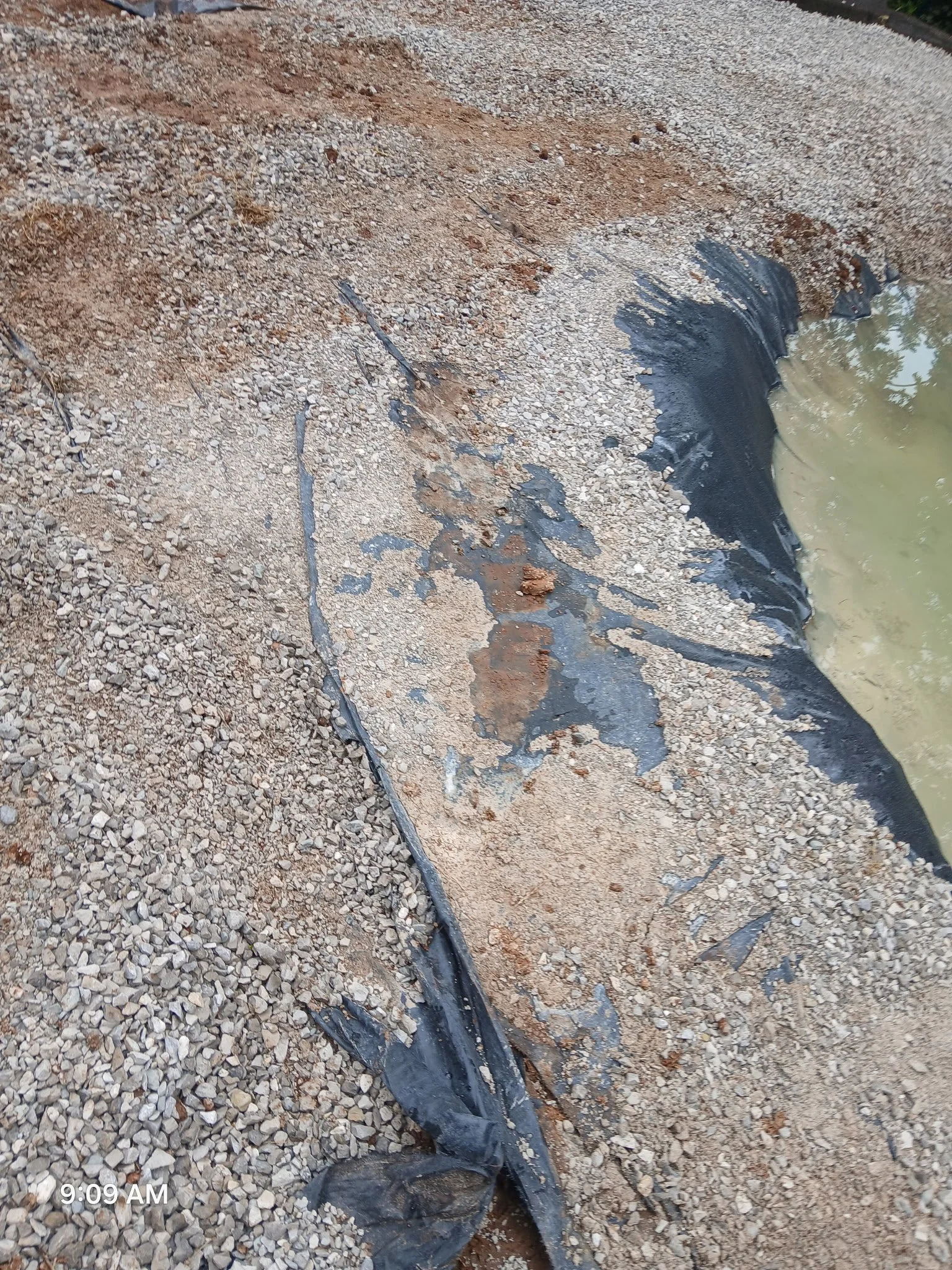 Close-up of a damaged pond liner on ground with gravel, with water visible on the right side.