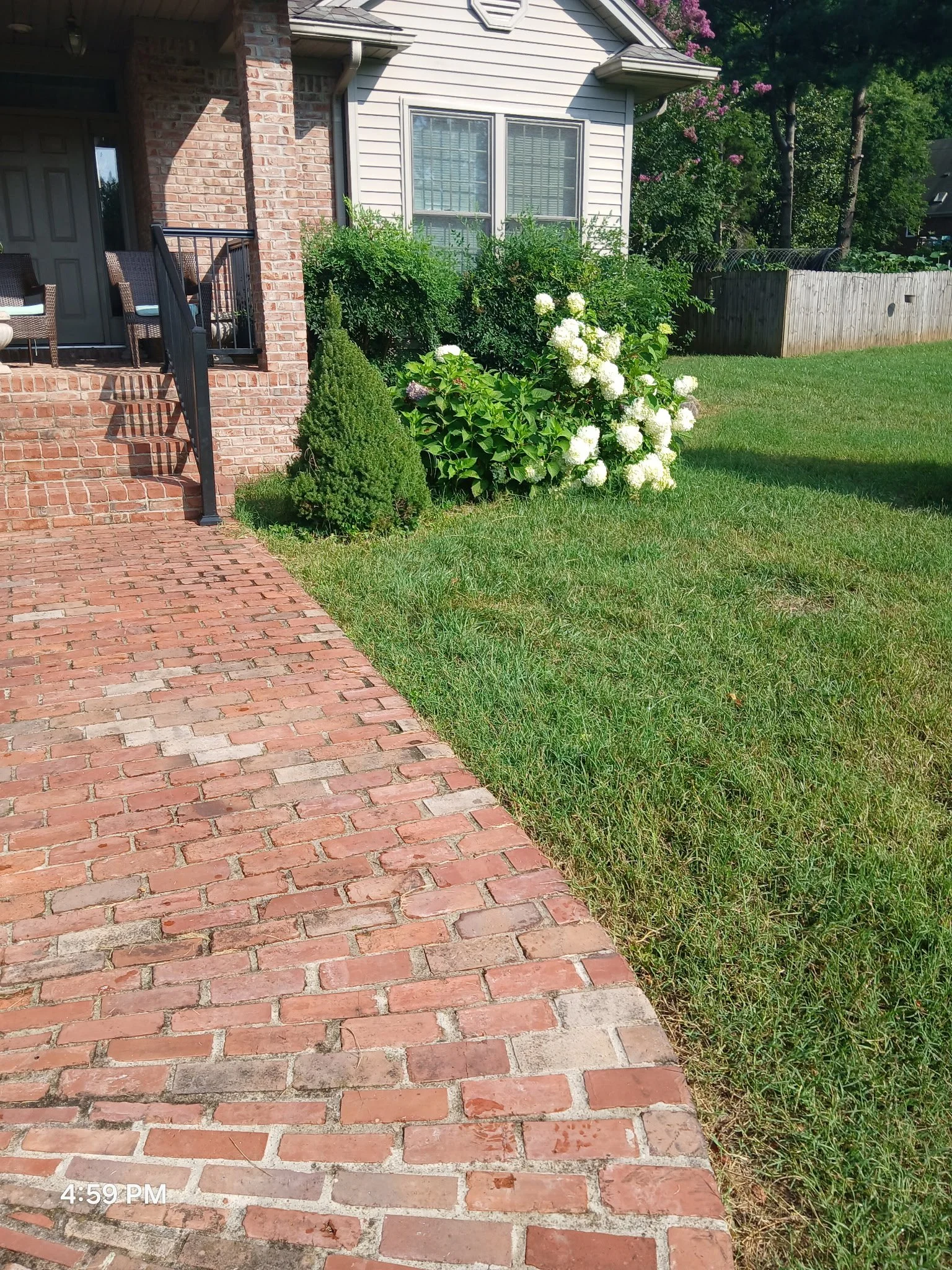 Brick walkway leading to a house with a small deck, surrounded by green grass and flowering bushes, with trees in the background.