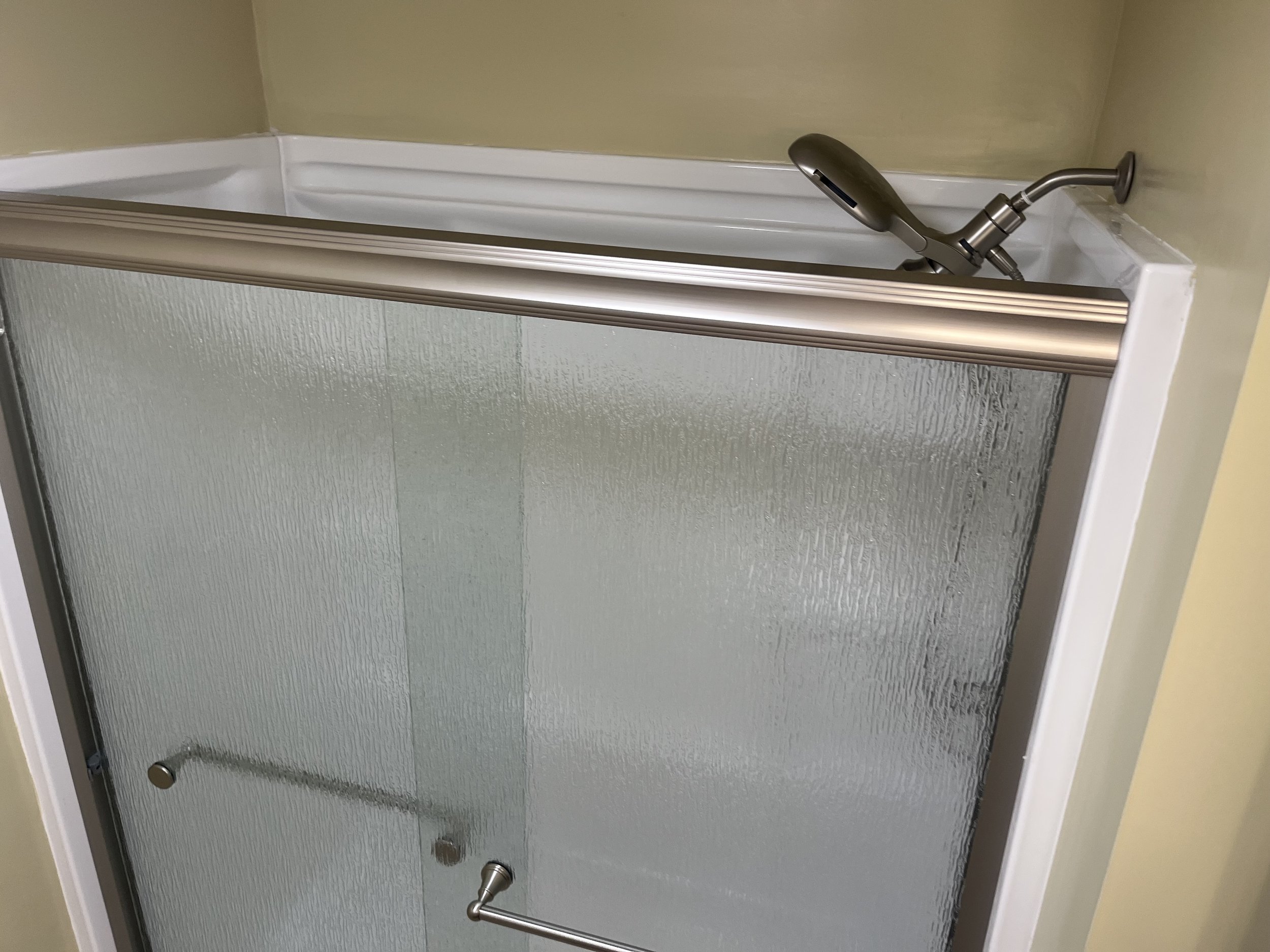 Shower with textured glass door, silver frame, and a handheld showerhead mounted on the wall inside.