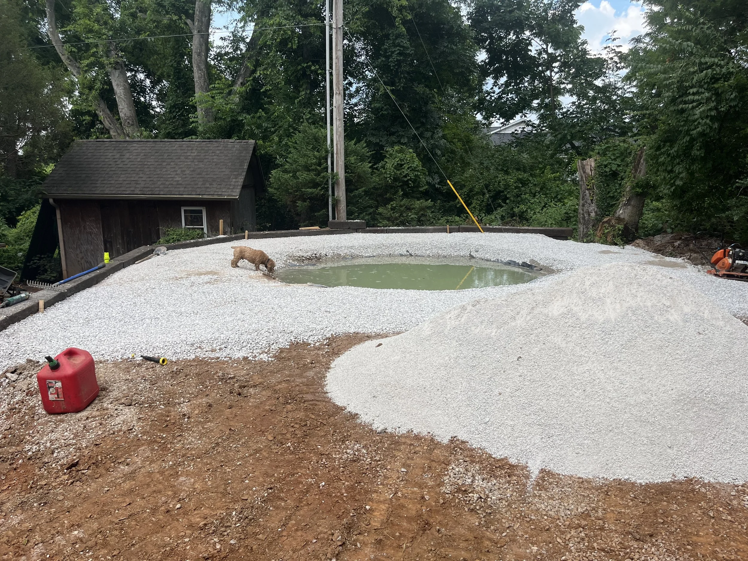 Under construction backyard with a small pond, gravel, and dirt, surrounded by trees, a shed, and construction equipment.