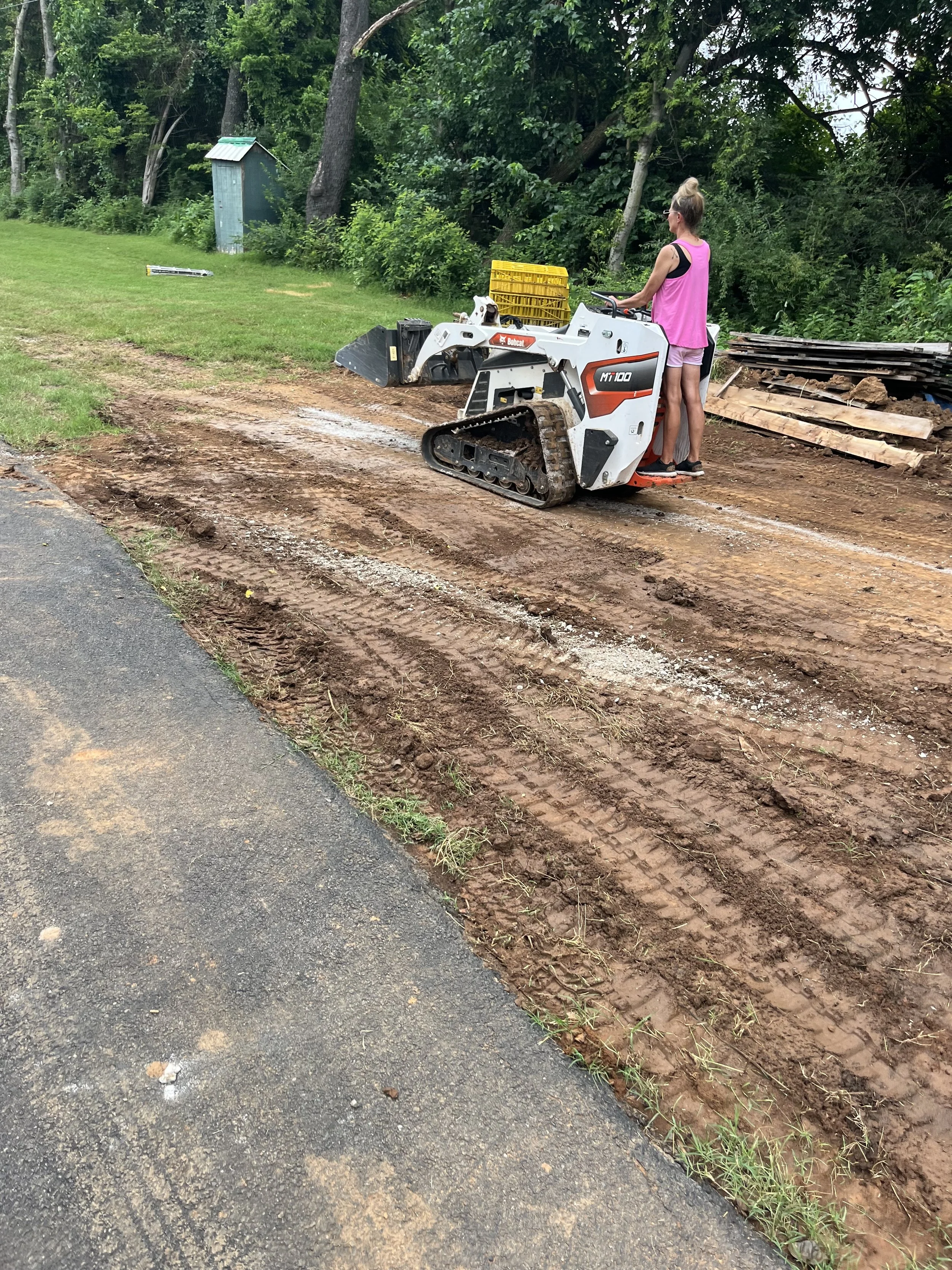 A woman operating a small bulldozer on a dirt path next to a paved road, with trees and vegetation in the background.