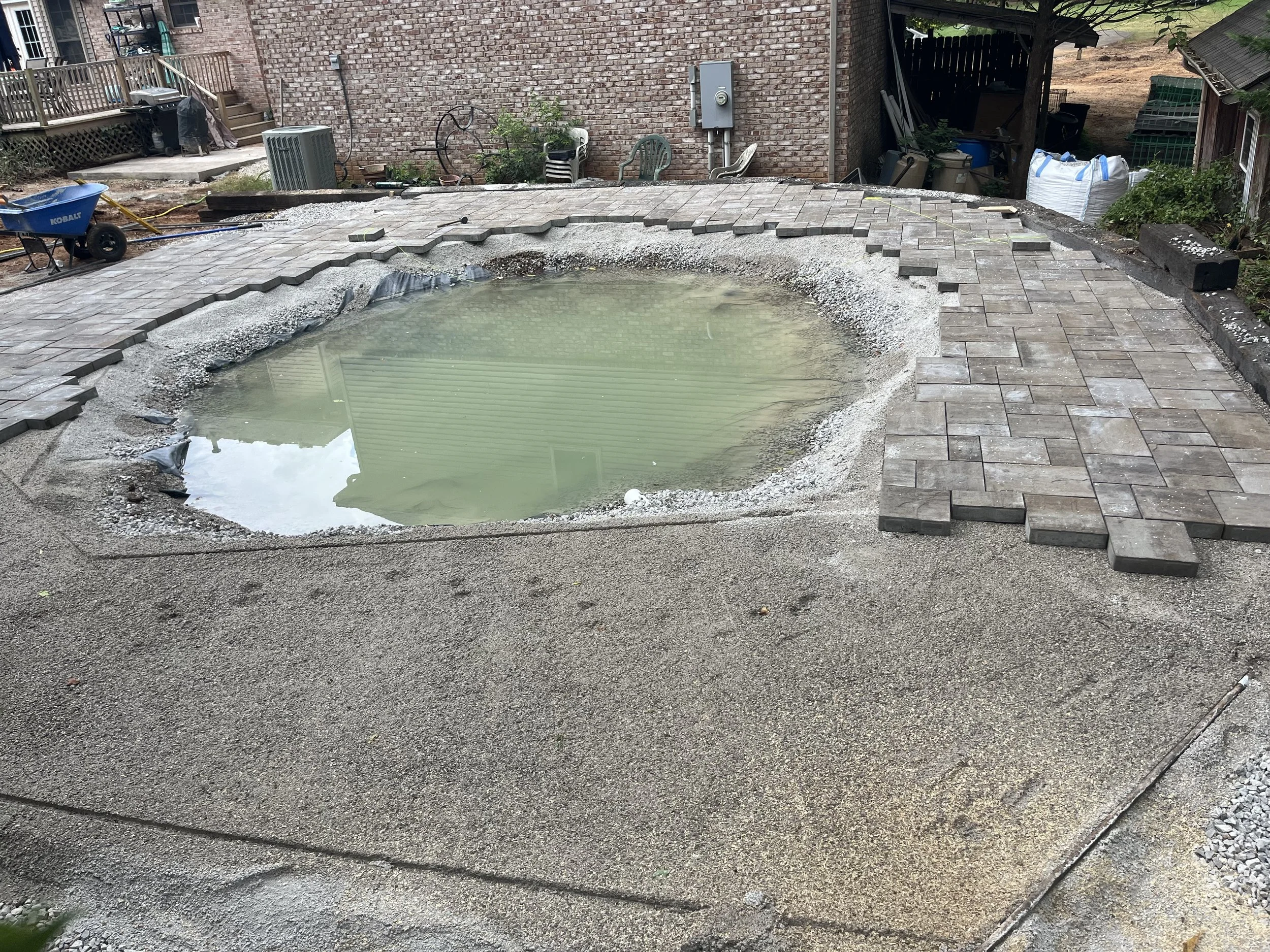 A backyard in the process of having a pool installed, with a partially filled and unfinished pool surrounded by paving stones and construction materials.