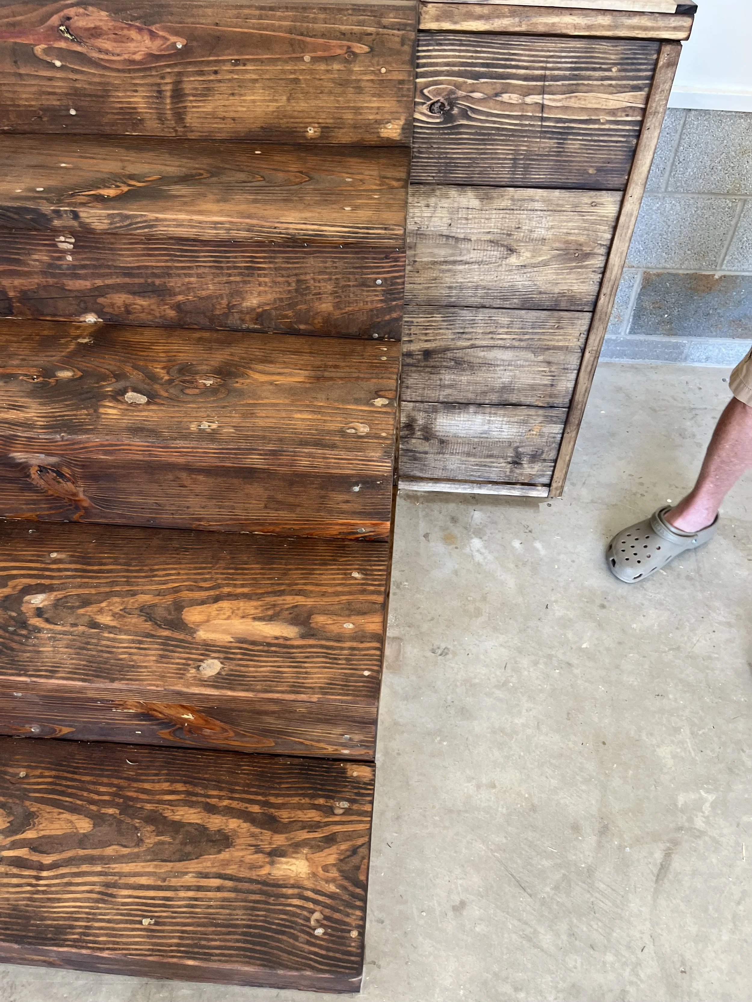 Close-up of wooden boards, some polished and stained dark on left, and others weathered and gray on right, with a person's foot wearing a gray croc visible in the lower right corner.
