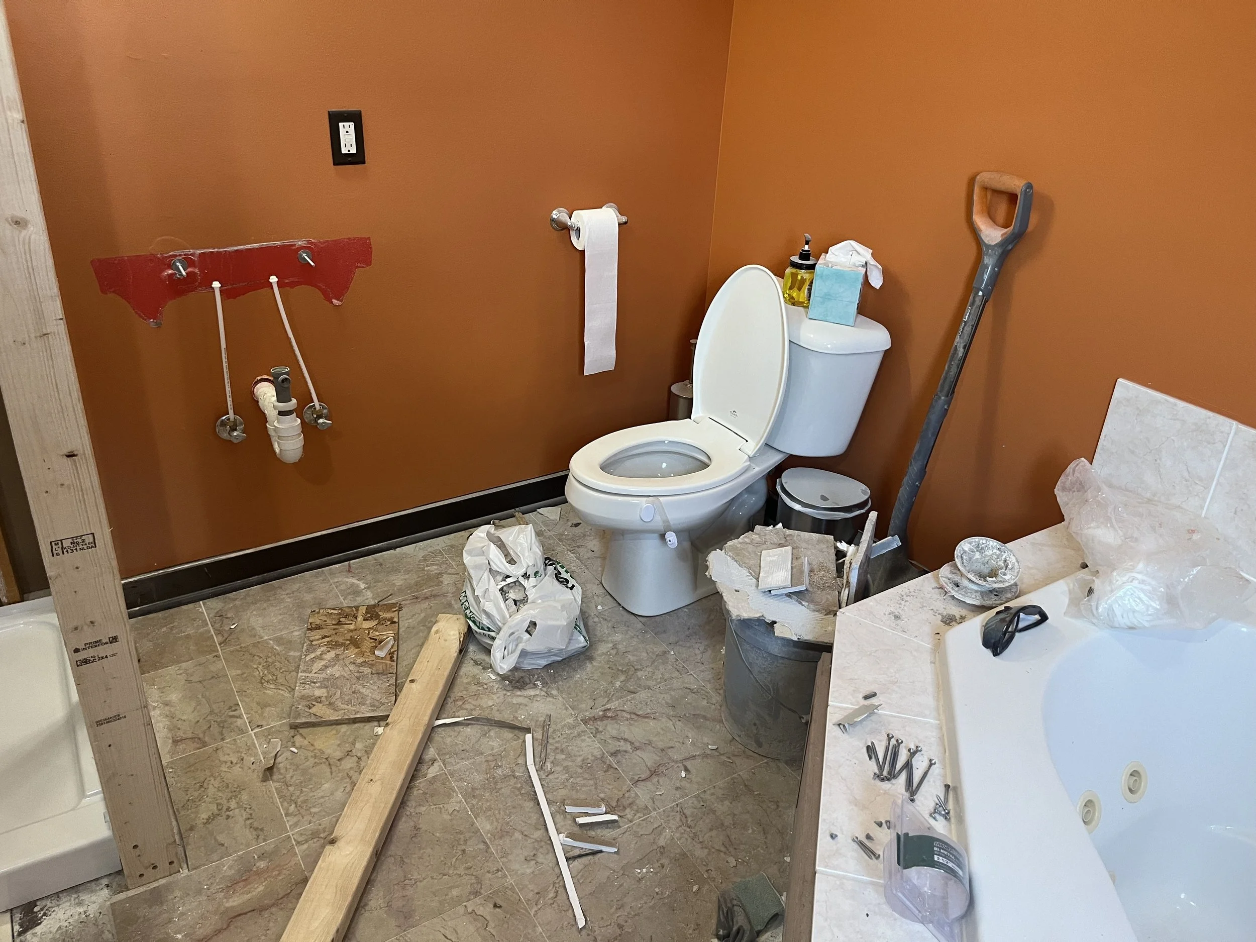 Bathroom under construction with tools, construction materials, and debris scattered around, including a toilet, a partially installed wall, and a bathtub.