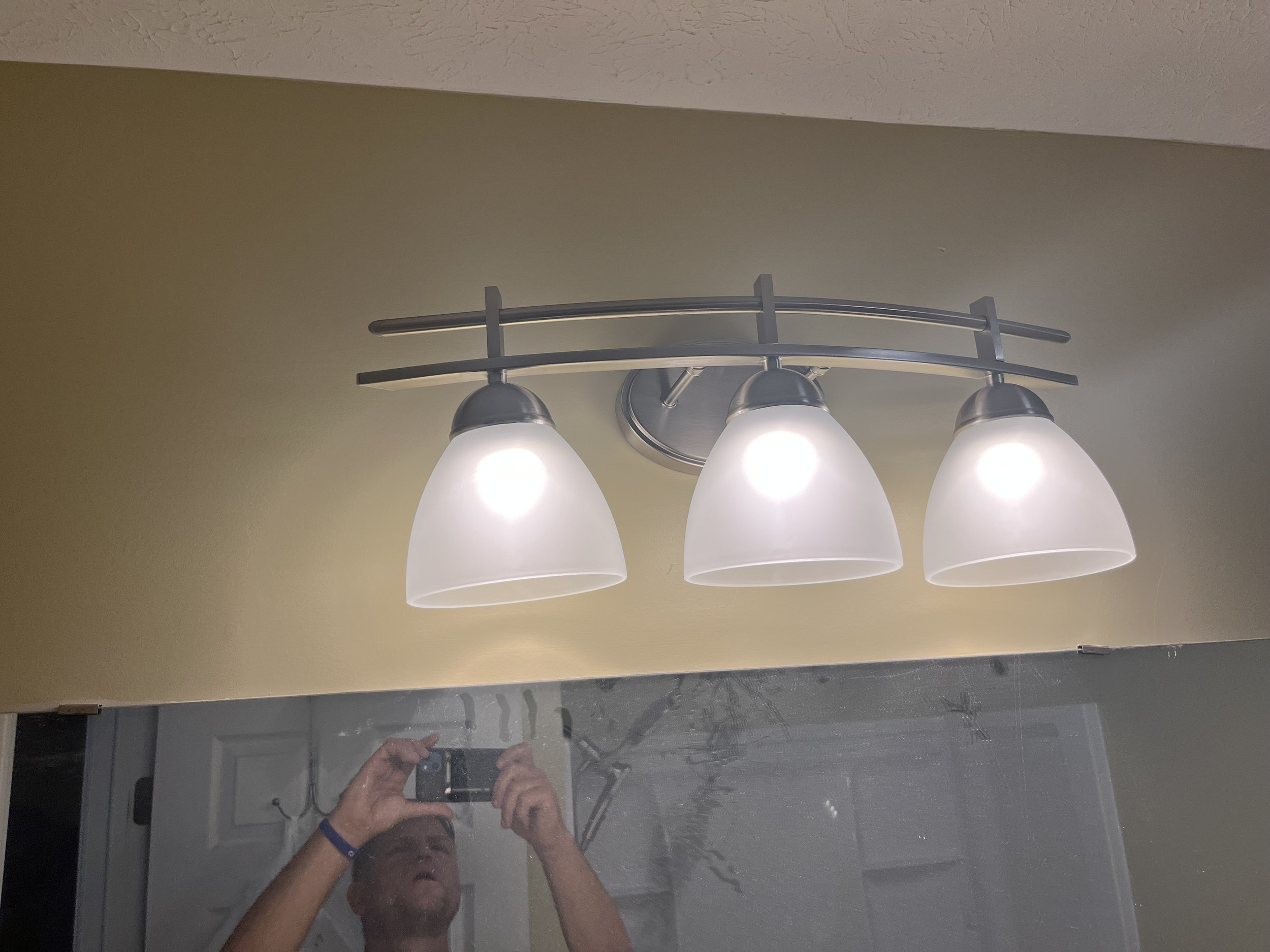 Three white frosted glass bathroom vanity light fixtures with silver metal mounts on a yellow-green wall, reflected in a mirror.