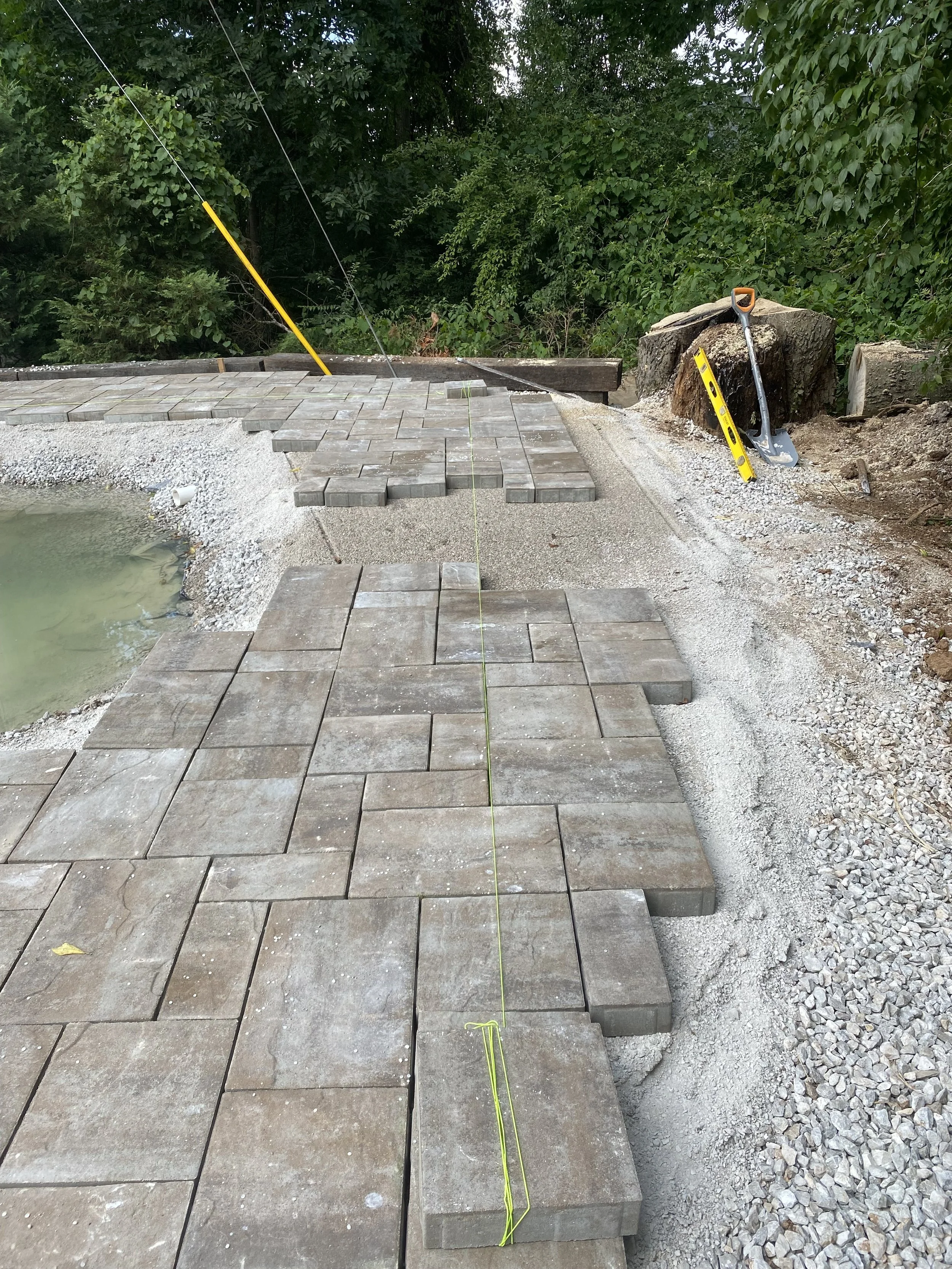Under construction of a stone patio or walkway with paving stones being laid and a level tool on top. A string line guides the placement of the stones, and a shovel rests against a large rock nearby. Surrounding trees and bushes are visible in the ba