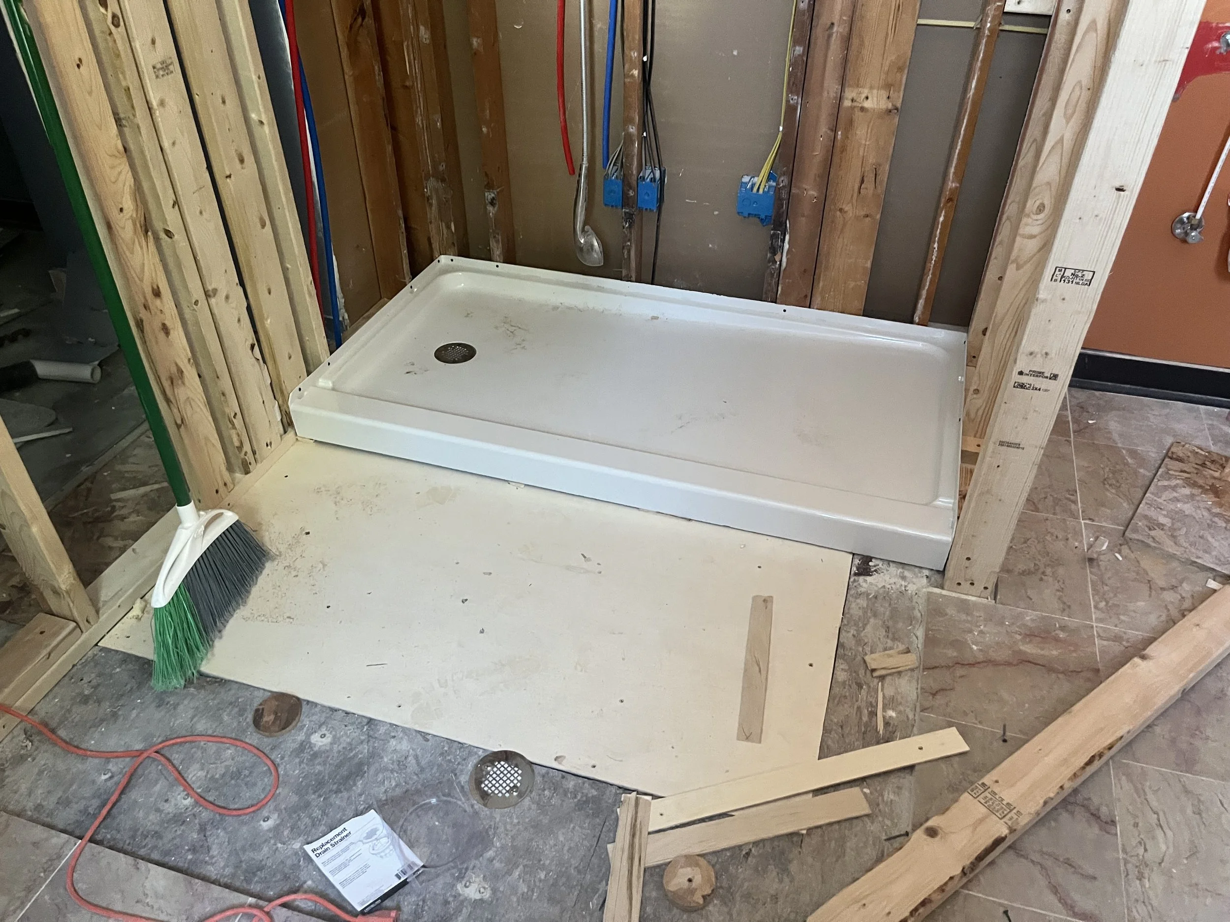 Construction site of a bathroom with a white shower base, exposed wooden framing, a broom, and some construction materials on the floor.