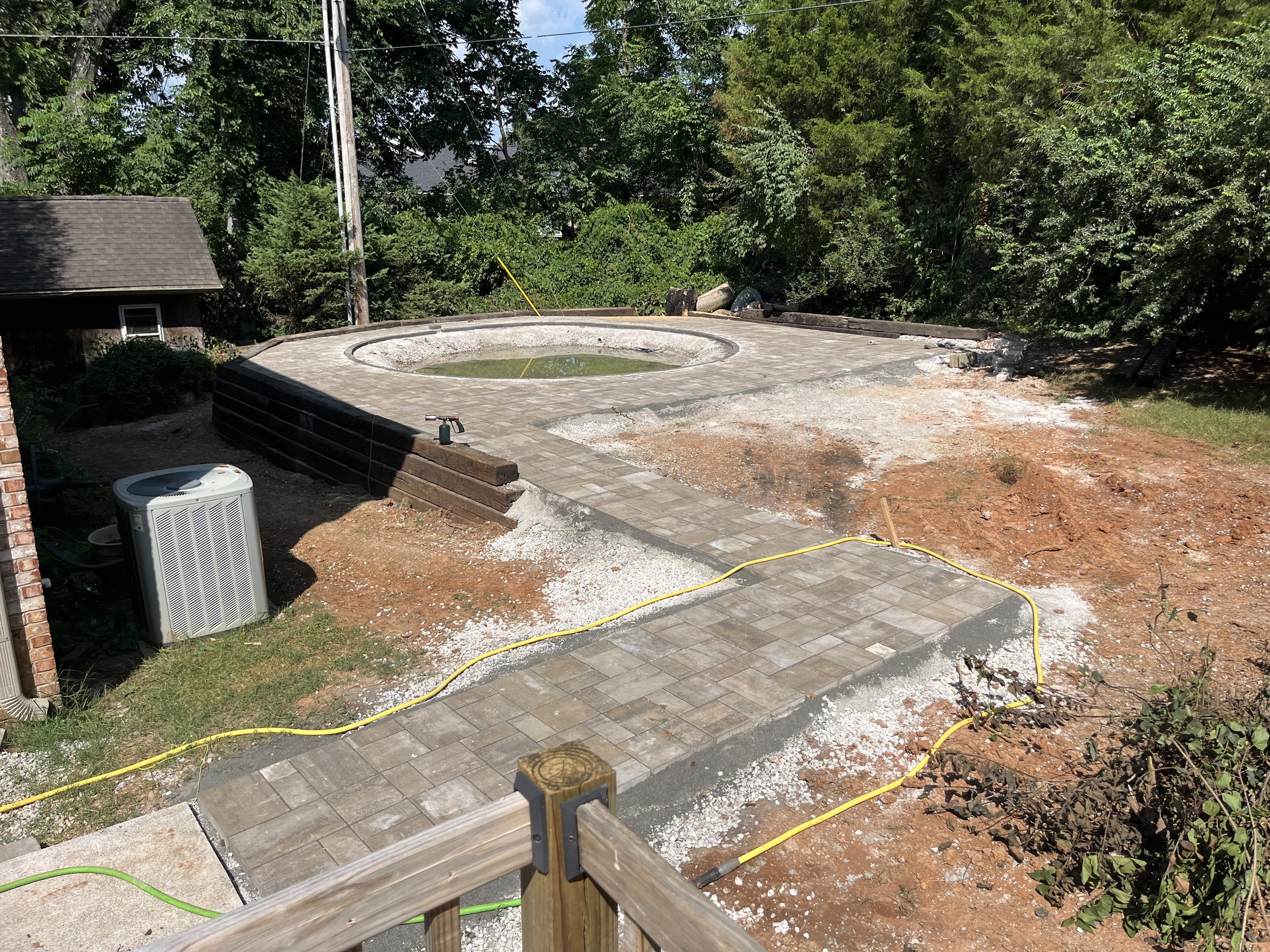 Construction site with partially built circular hot tub or small pool, paving stones, and construction tools. Surrounding area has dirt, gravel, and some vegetation with trees in the background.