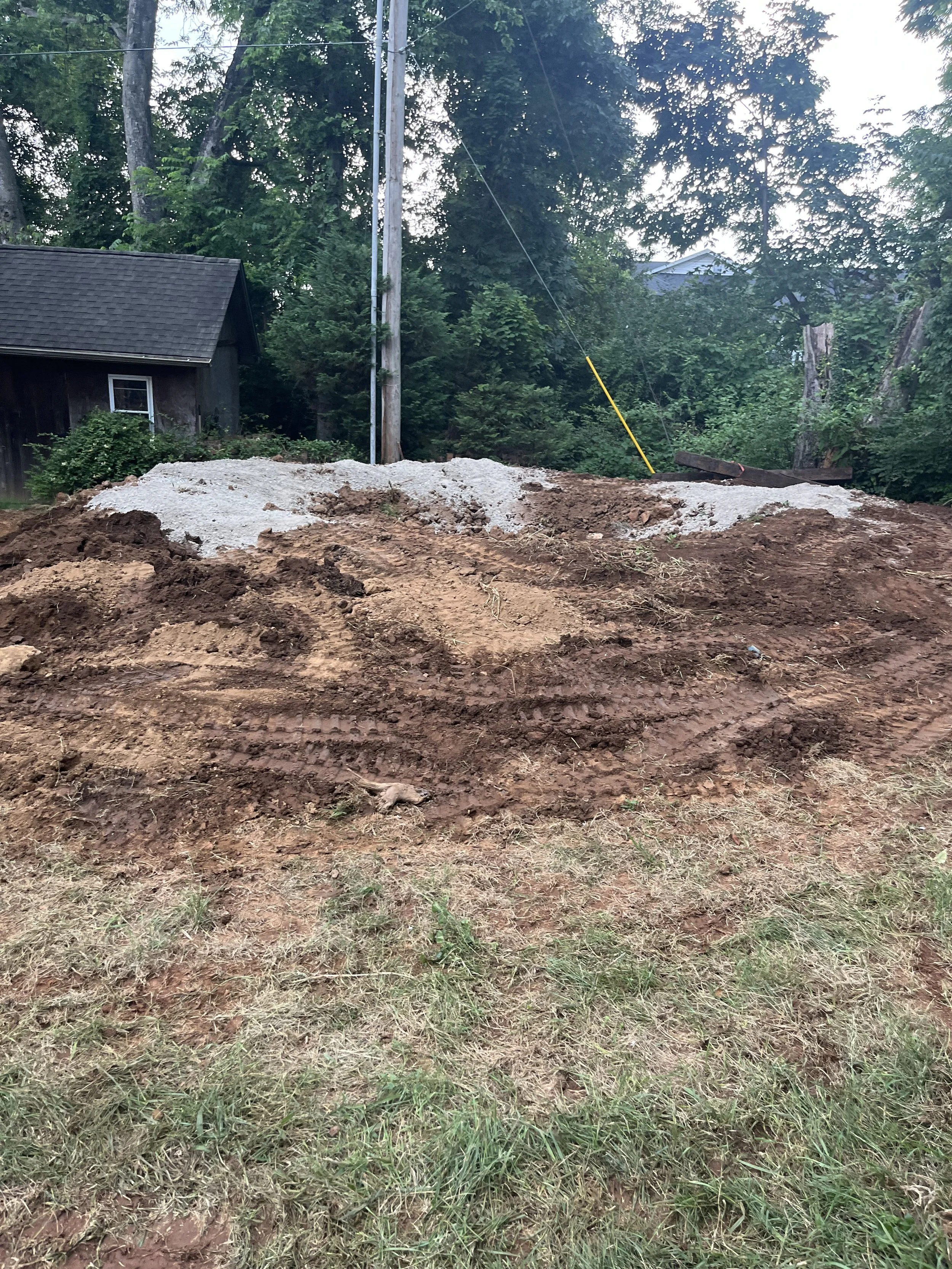 A construction site with dirt and gravel, with a slope of exposed earth, nearby trees, and a utility pole in the background.