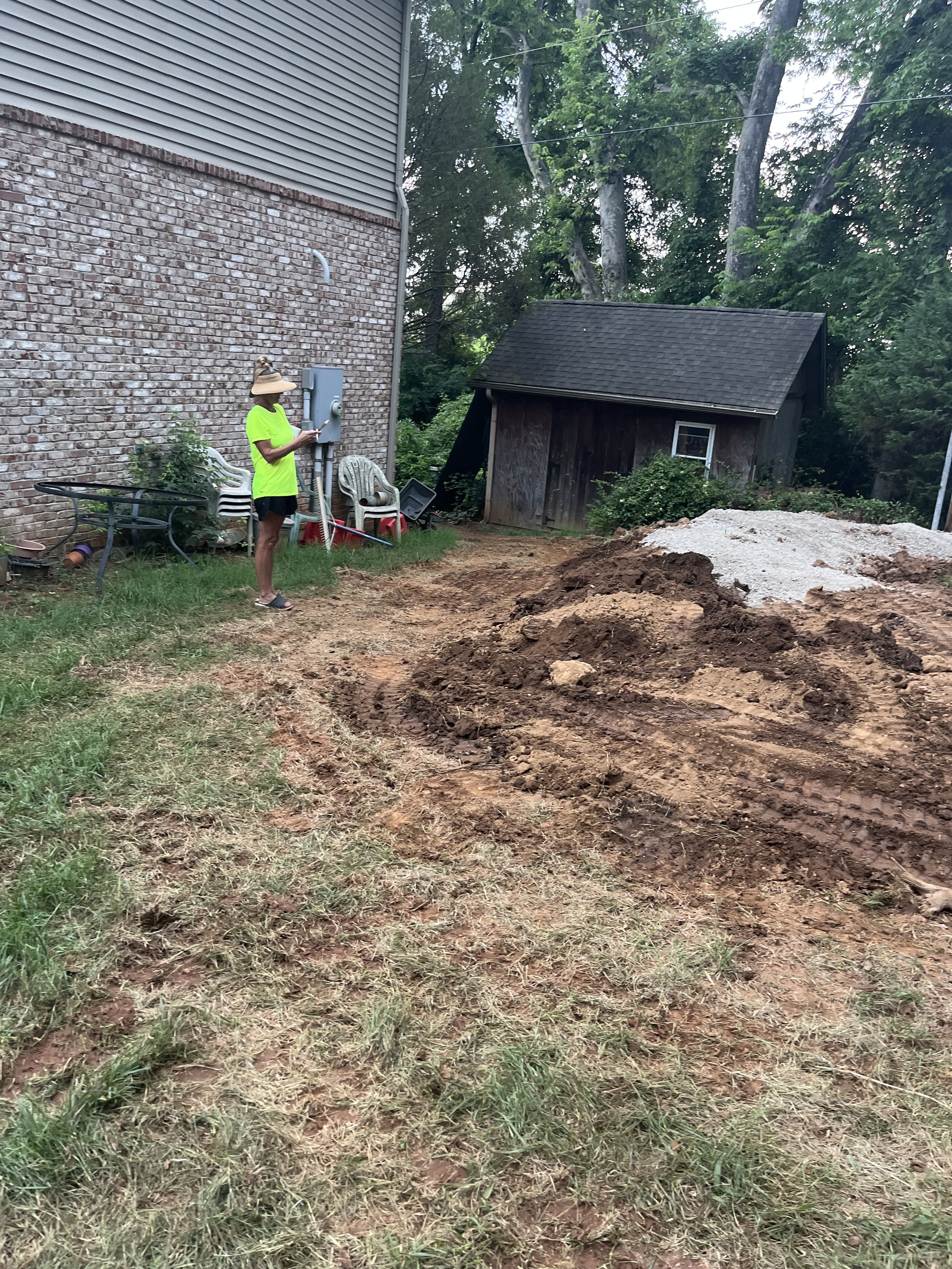 Person standing near a brick house, operating a control panel, with a pile of dirt and a small shed in the background.