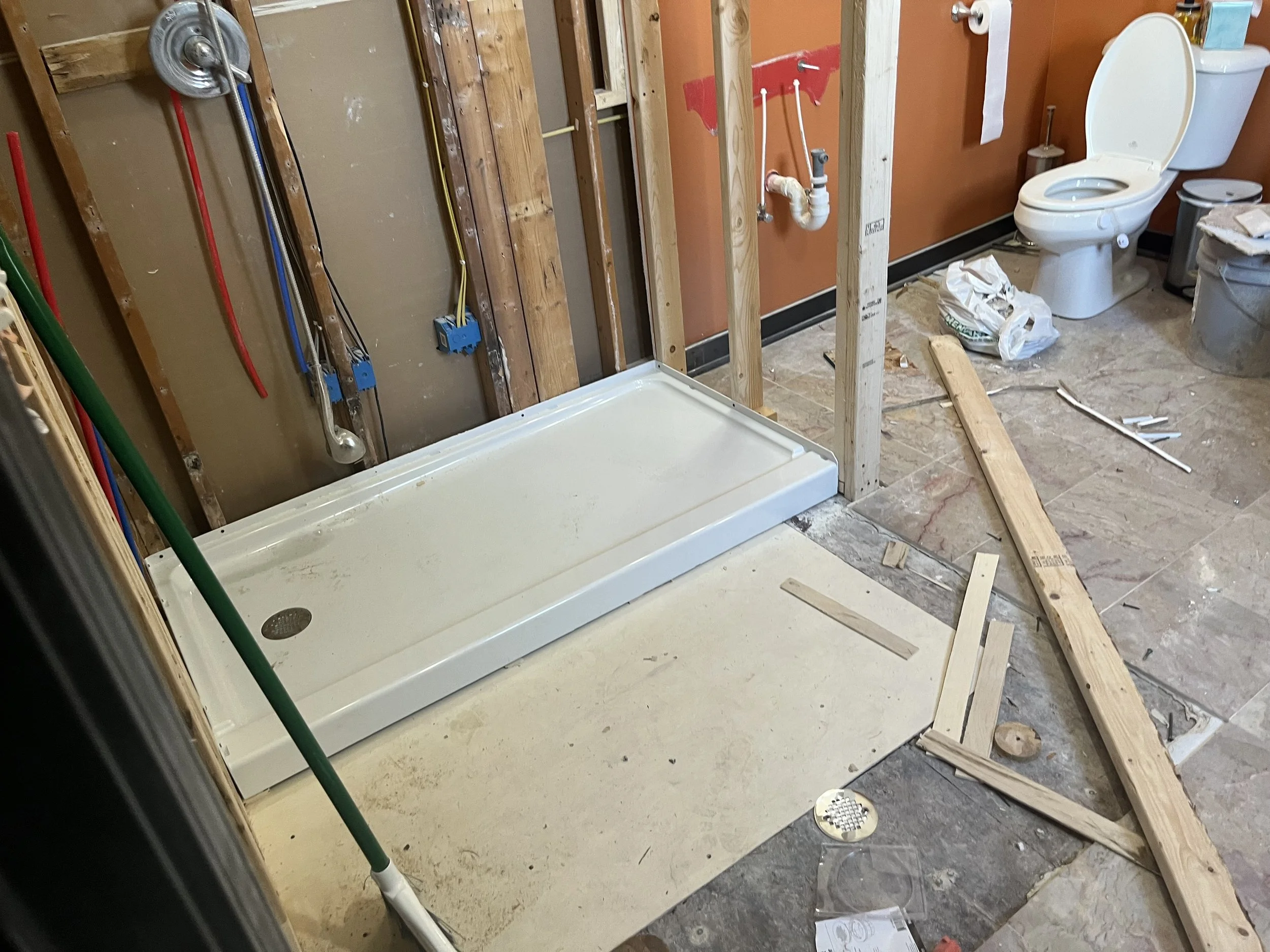 Bathroom renovation in progress with a shower base installed, a toilet in place, exposed wall studs, and construction tools and materials scattered on the floor.