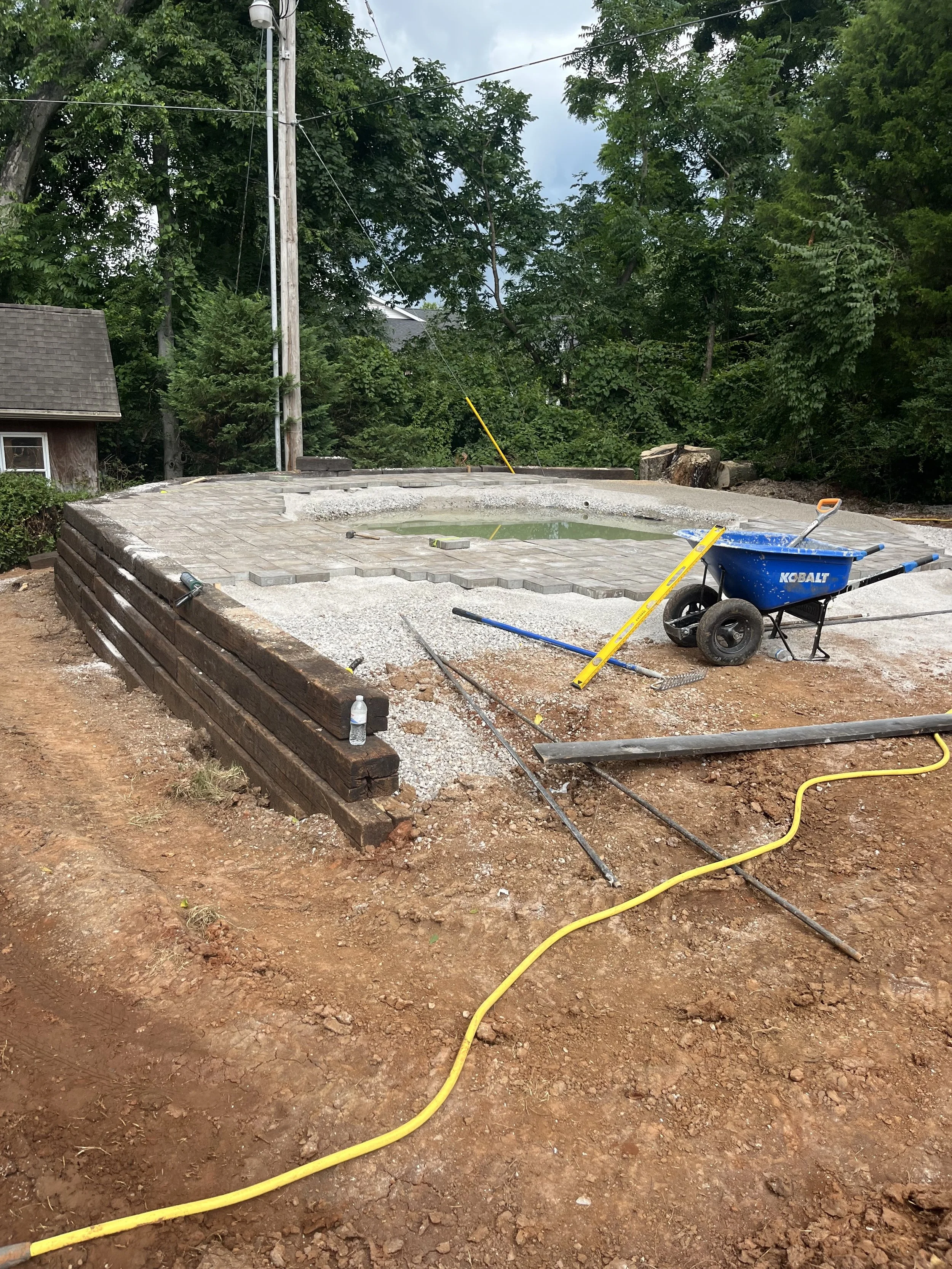 Under construction site with a kidney-shaped pool or pond, surrounded by paving stones. Building materials and tools are scattered around, including a blue wheelbarrow, leveling tools, and a yellow hose on the bare dirt ground. Trees and a small buil