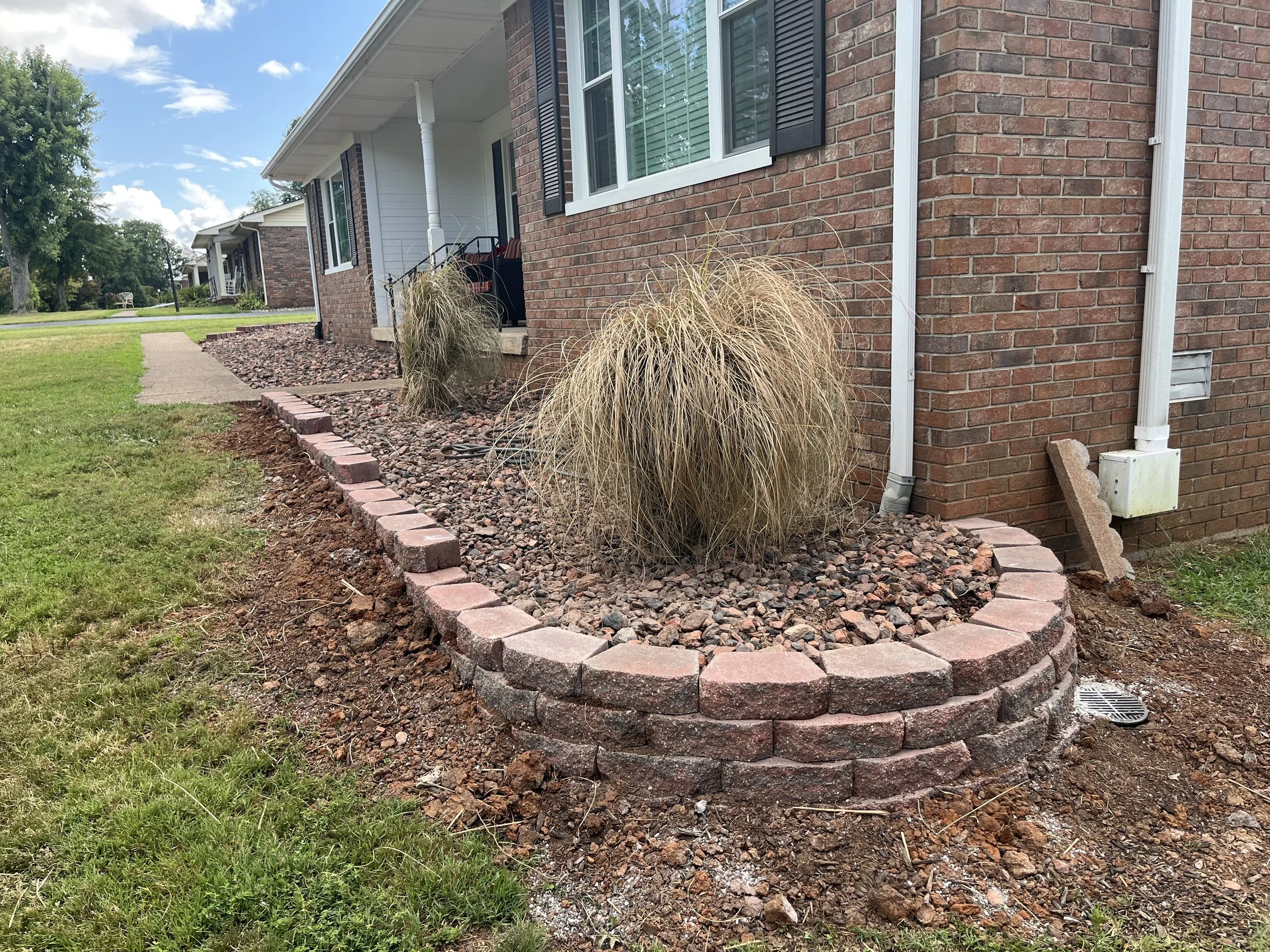Landscape view of a brick house with a landscaped garden bed featuring decorative grasses, a brick border, and gravel mulch. The house has white and black window shutters and gutters, with a sidewalk leading away from the garden bed. Green lawn and t