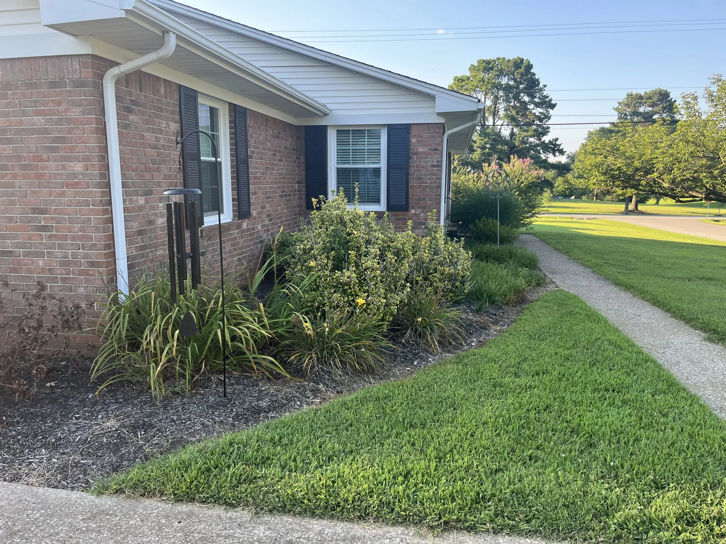 A brick house with two windows and black shutters, a white gutter, and a landscaped front yard with green grass, plants, and a small garden bed. A sidewalk runs next to the yard, with trees and street visible in the background.