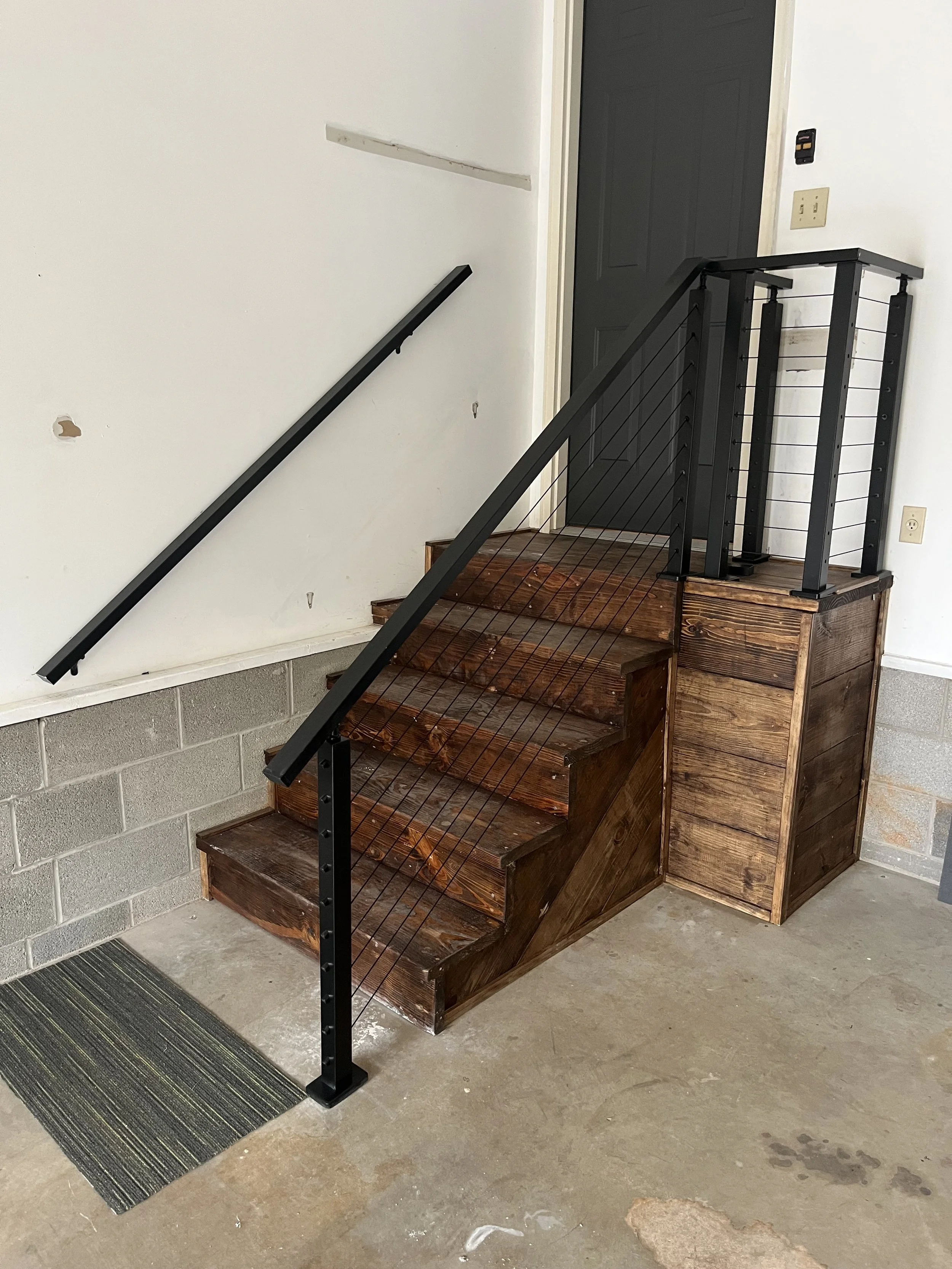 A small staircase with dark wood steps and a black metal railing leading up to a black door. The staircase is built with a wooden box-like structure. The area around the stairs is a concrete floor with some minor stains and a small striped doormat at