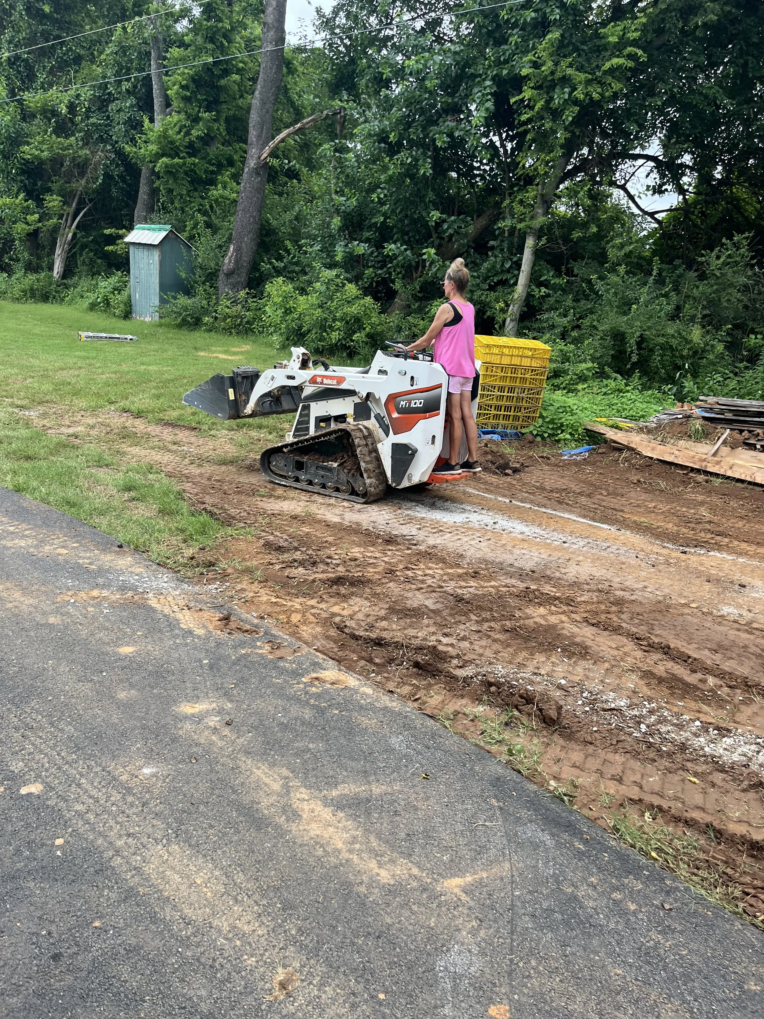 A woman operating a small compact tracked excavator on a dirt roadside next to grass and trees, with a yellow crate behind her.