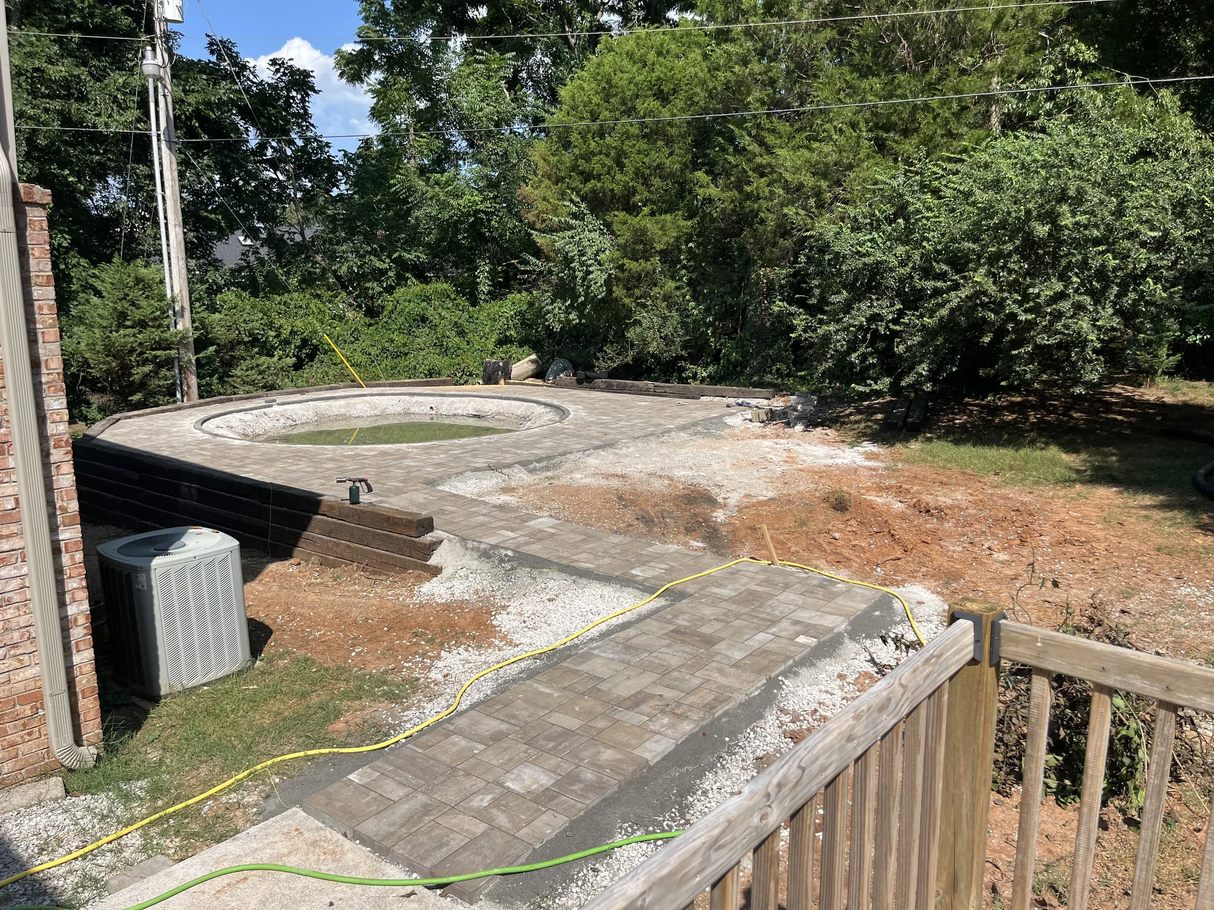 Backyard with a newly paved patio area around a circular pool under construction, surrounded by trees and vegetation, with construction tools and equipment visible.