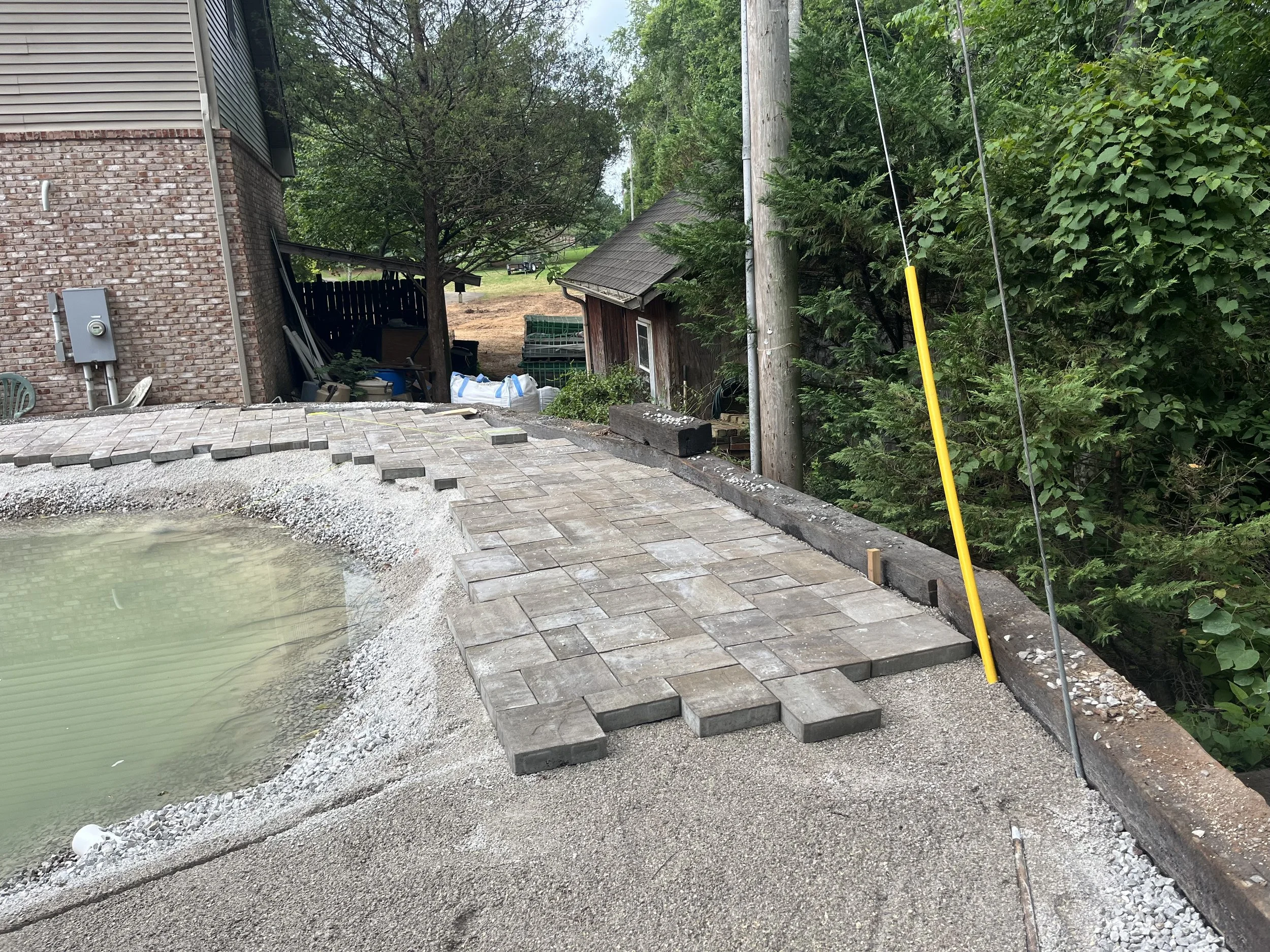 Creating a paved pathway along a residential backyard with the pool on the left and greenery on the right, with construction tools and materials present.