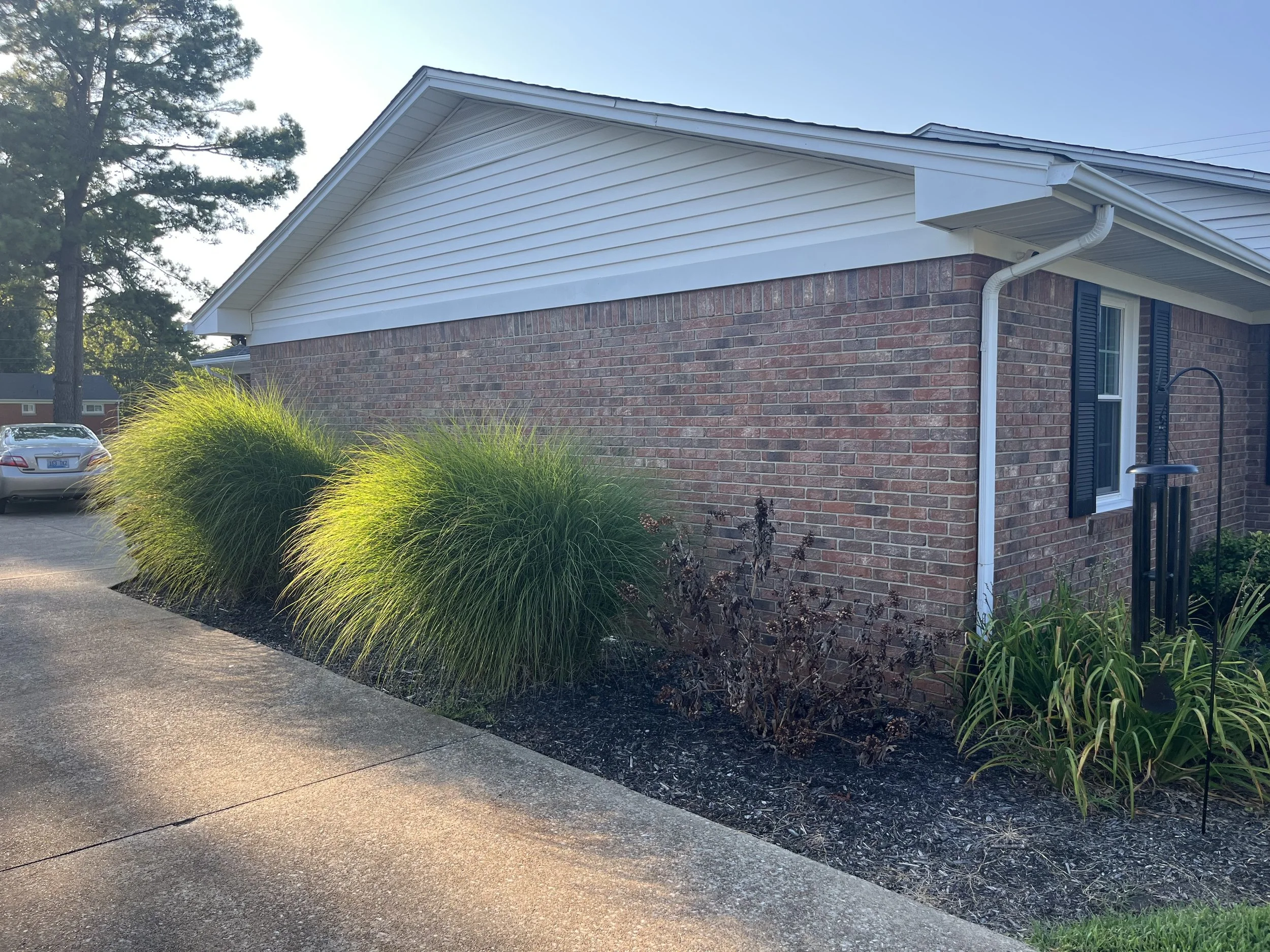 Exterior of a brick house with white siding on the roof, two windows with black shutters, a white gutter system, and landscaped bushes and plants in front.