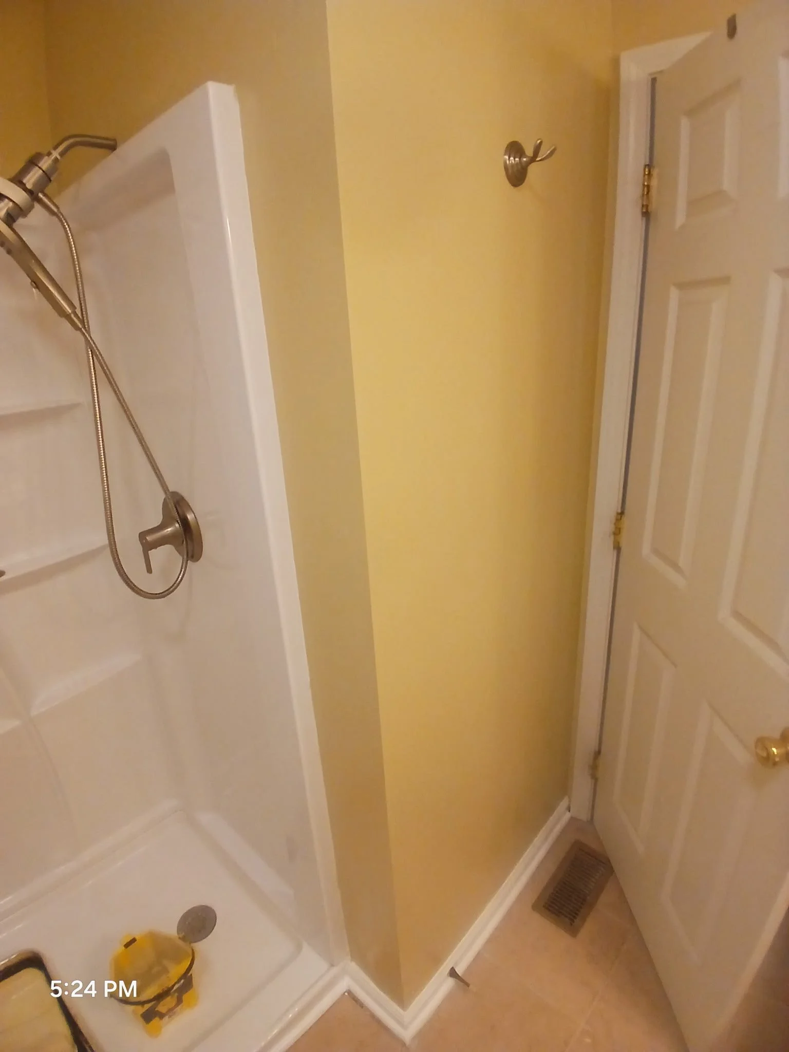 Corner of a small bathroom showing a shower stall with a handheld showerhead, a white door with a gold doorknob, a yellow wall, and a small wall hook.