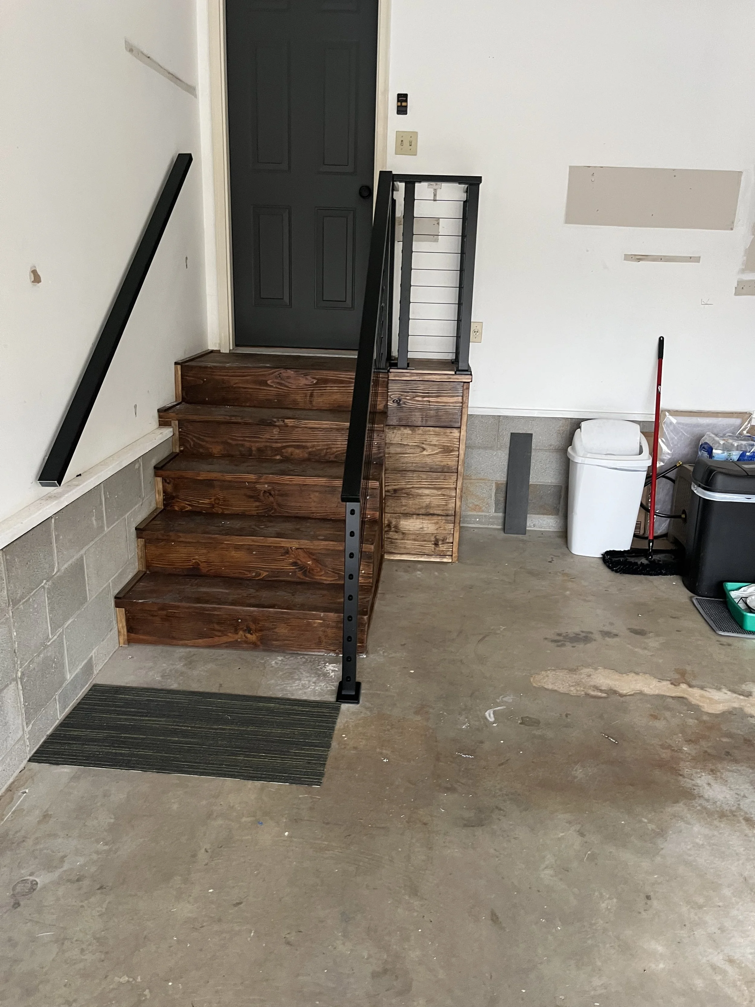 A small wooden staircase with four steps and a black metal railing leading to a dark gray door in a garage or basement area. To the right, there is a white trash can, a red broom, and some storage bins.