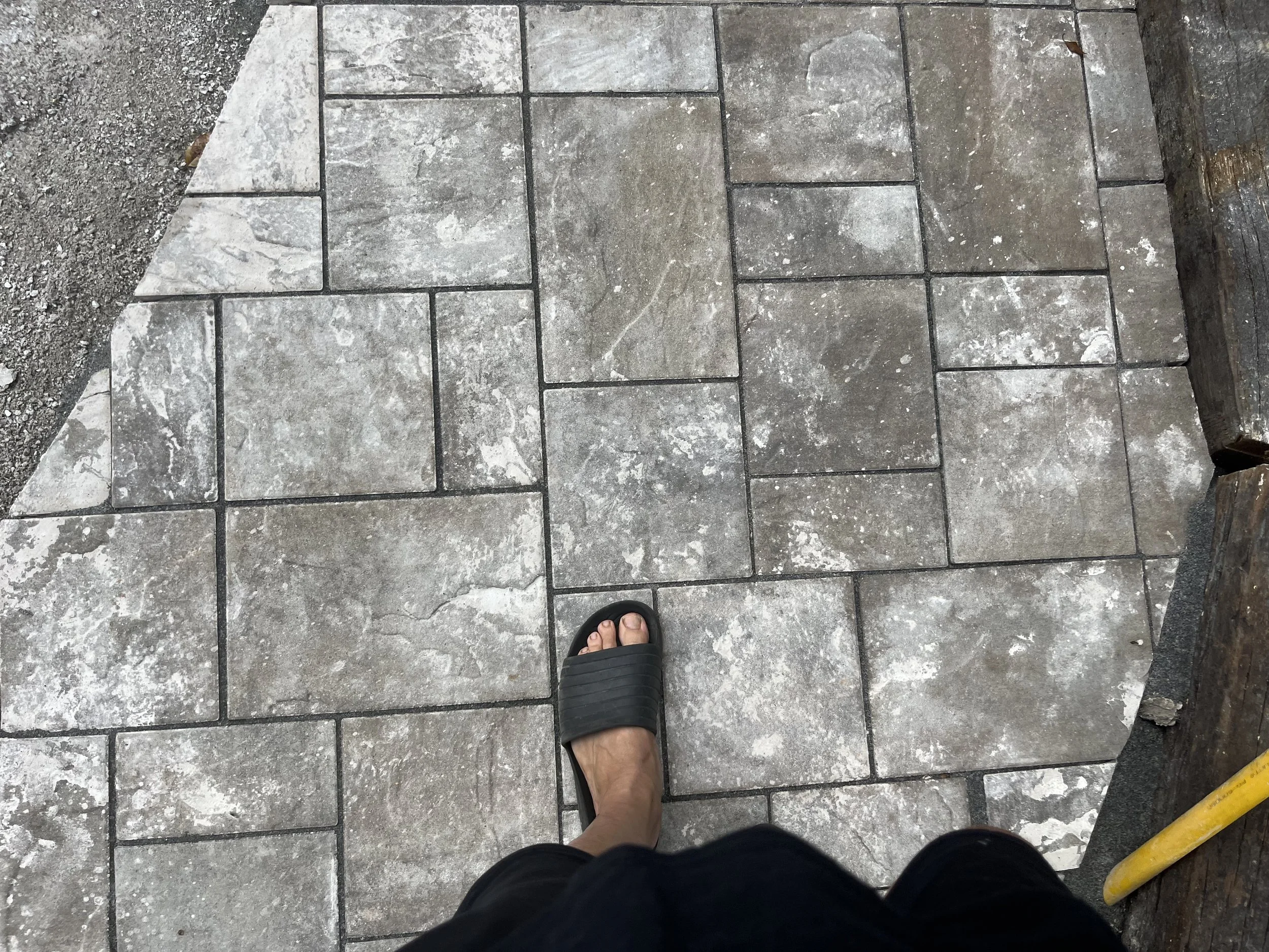 A person's foot in a black sandal stepping on a stone pavement with rectangular and square gray tiles, with part of a yellow pole and a wooden edge visible.