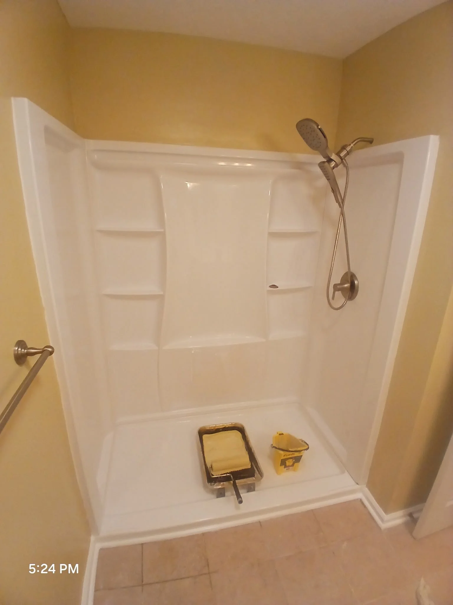 Empty white shower with a handheld showerhead, built-in shelves, and a beige tiled floor. A paint roller tray and a can of paint are on the shower floor, indicating ongoing renovation.