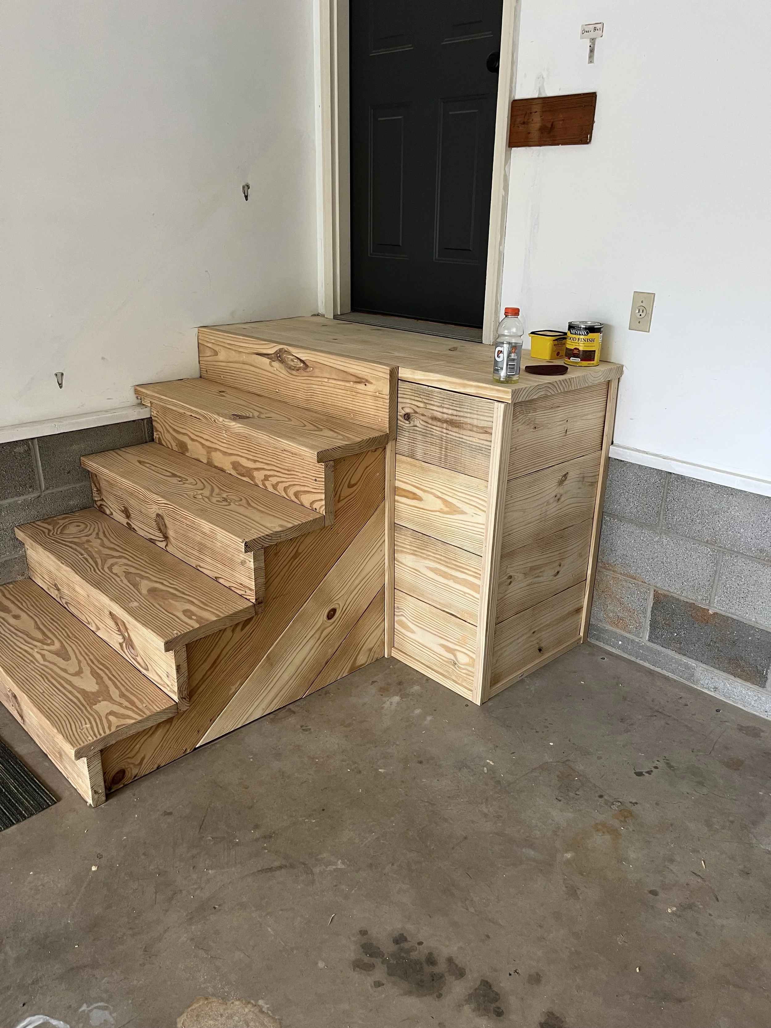 Wooden stairs leading to a small elevated landing near a black door, with some tools and supplies on top of the landing.