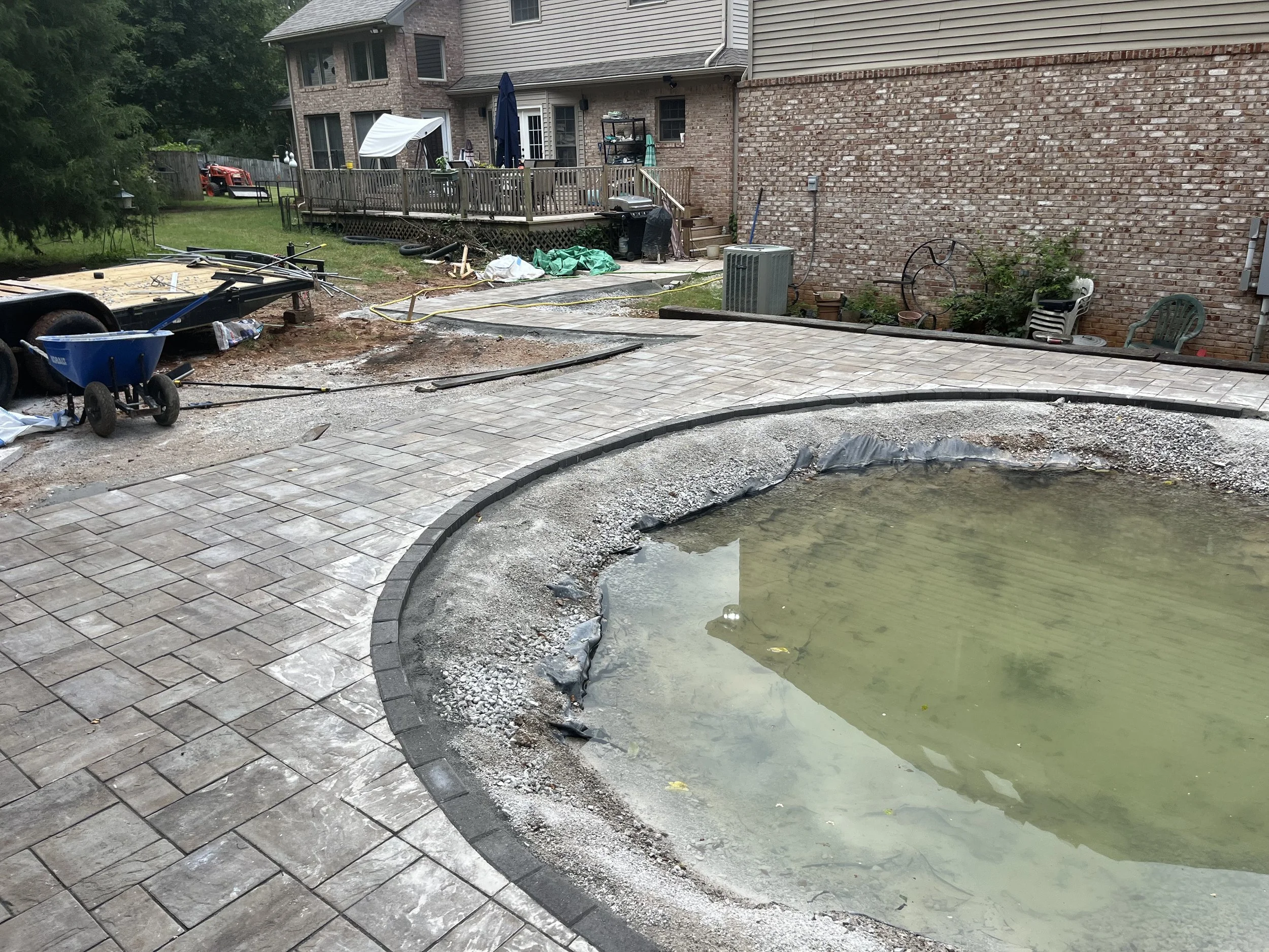 Backyard pool renovation under construction with a partially filled concrete pool, paver stones being installed around it, construction tools, and materials scattered in the area.