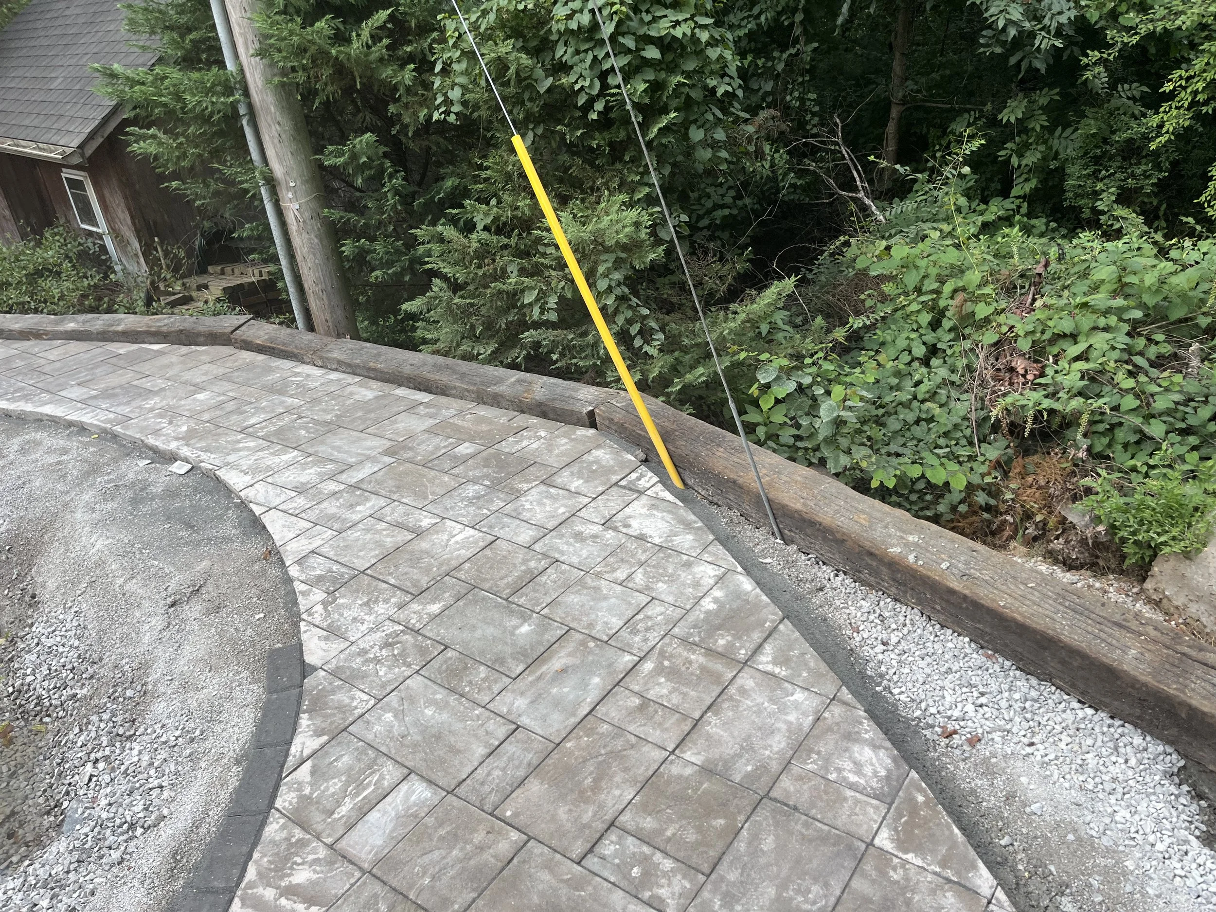 Newly paved outdoor pathway with brick pattern, bordered by gravel and a wooden beam, next to dense green foliage and trees.