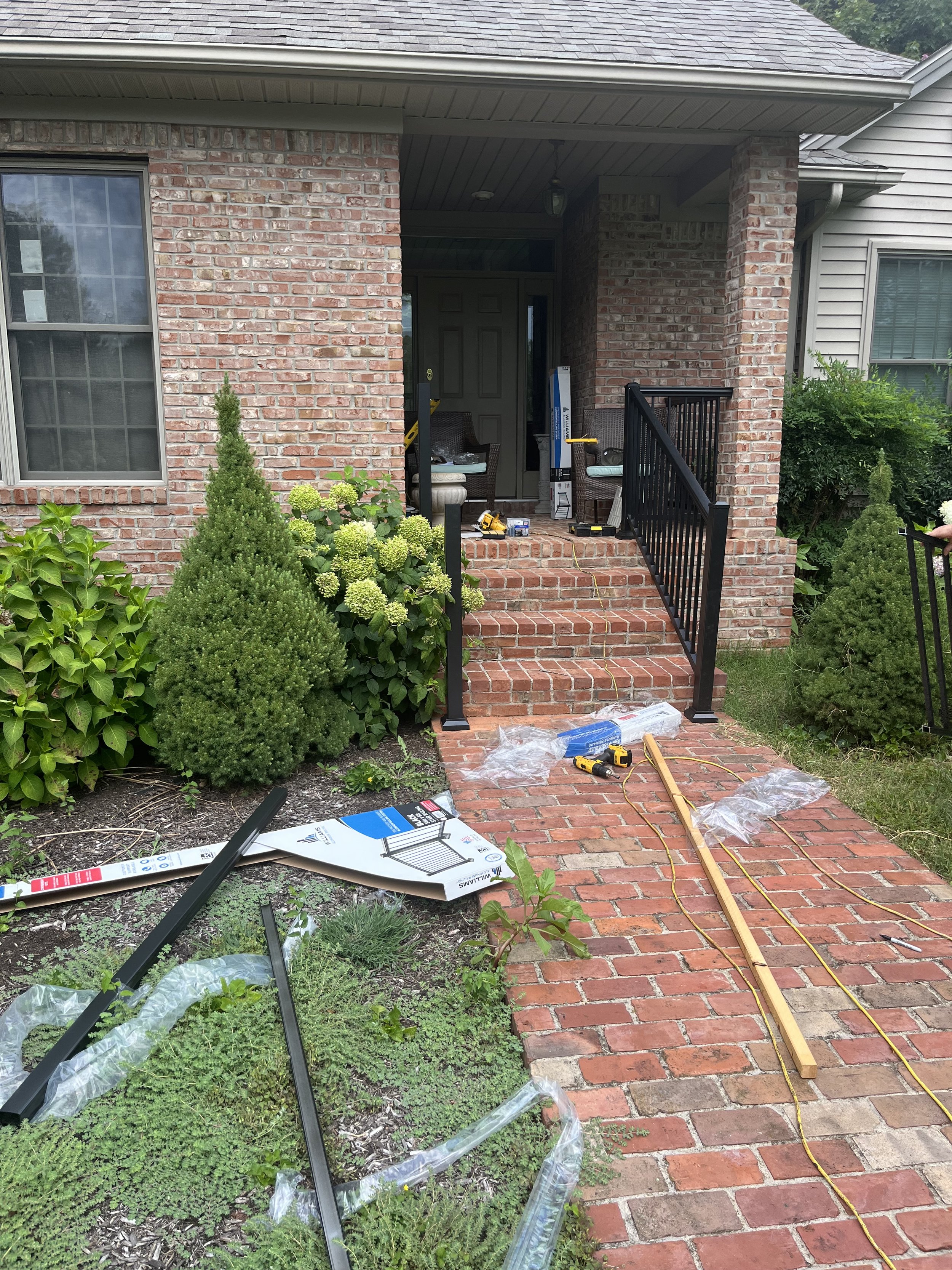 Front porch of a house under construction with brick steps, black railing, garden bushes, and tools on the steps and walkway.