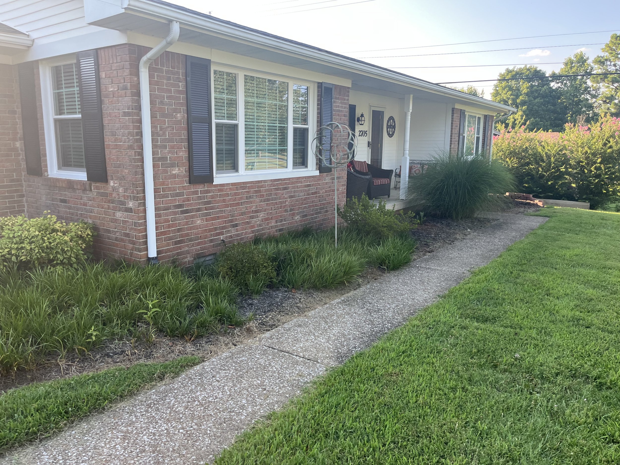 Front yard view of a single-story house with brick and white siding, black window shutters, a concrete walkway, green grass, and plants.