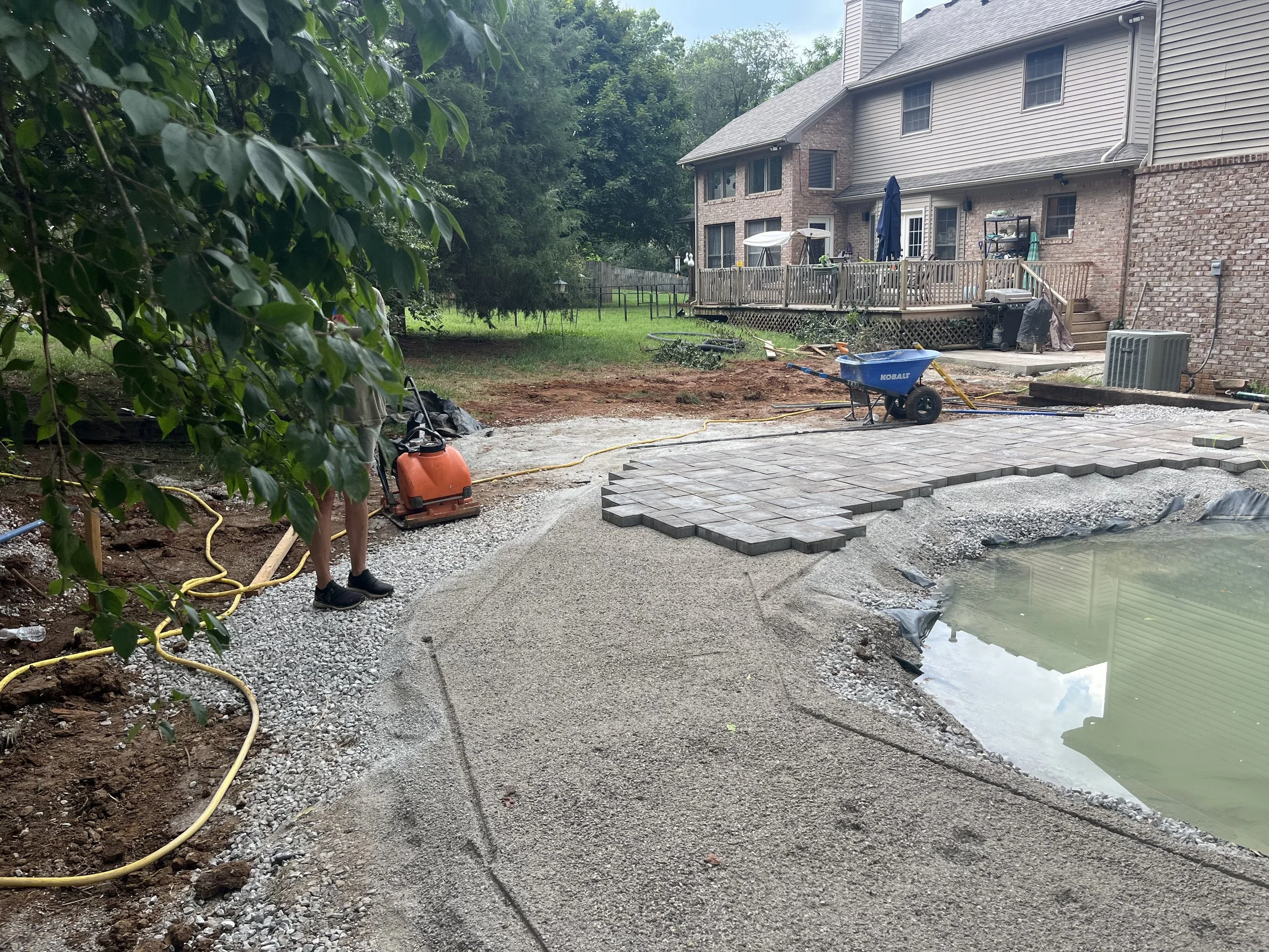 Backyard area under renovation with a partially paved stone patio, a small pond, and construction tools like a wheelbarrow and power equipment. Part of a house with a deck is visible in the background.