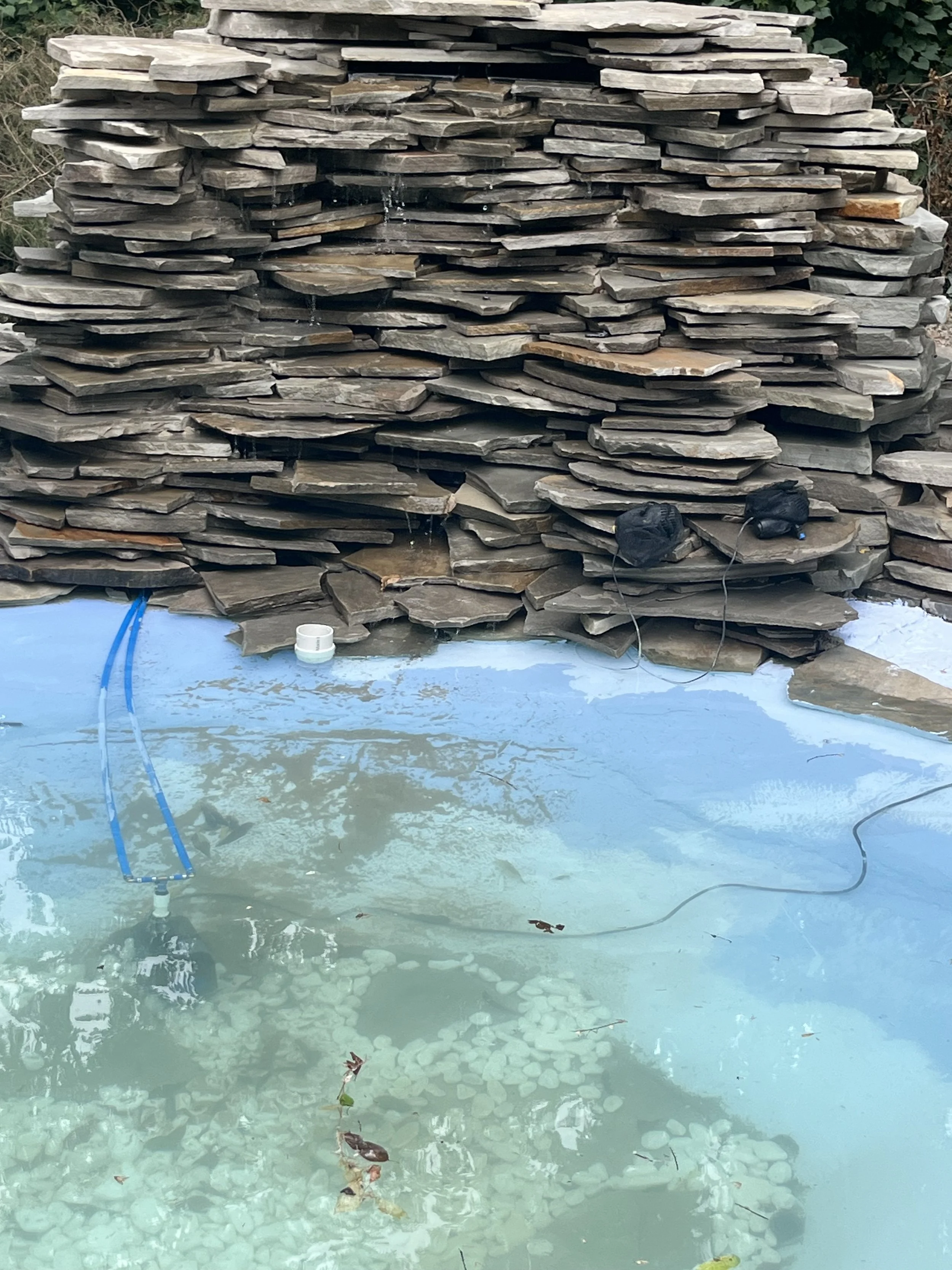 A backyard pond with a stone waterfall feature, blue tubing, and a white pipe visible in the water, with some fallen leaves floating on the surface.