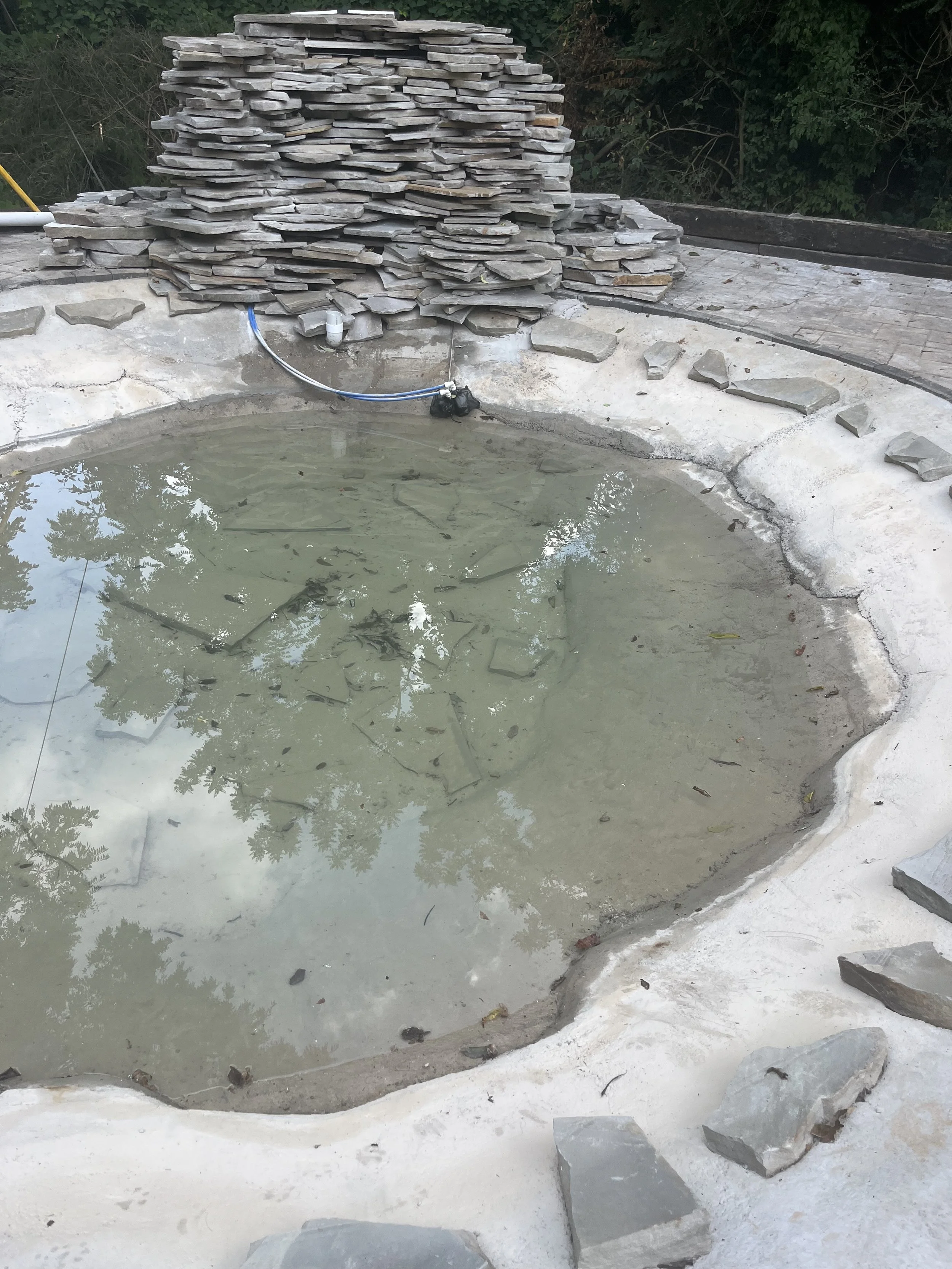 A small, partially filled stone hot tub with murky water. There is a stone wall with a pipe at one end, and a pile of flat stones stacked on top of the wall. Surrounding the hot tub are some loose stones and green bushes.