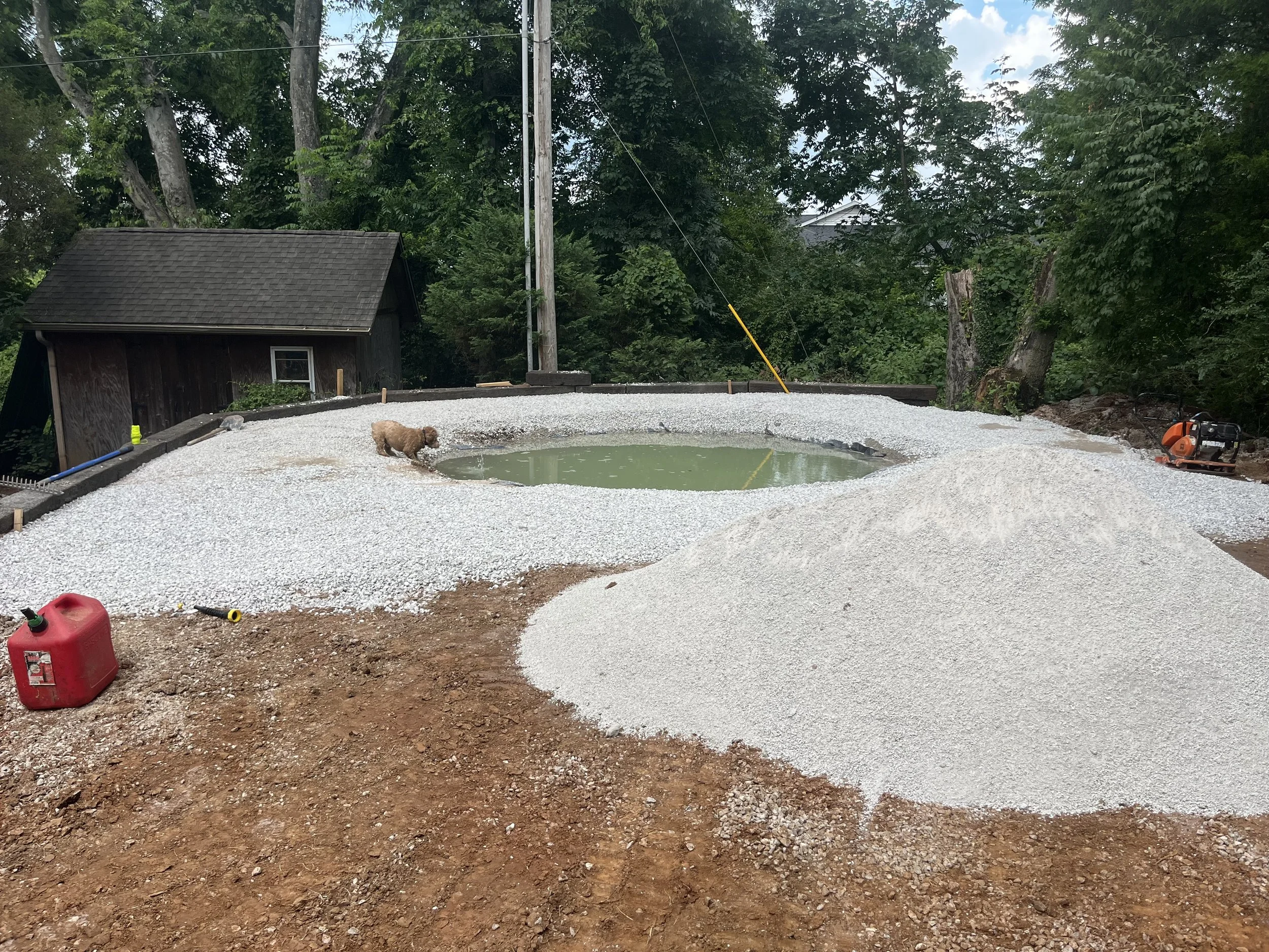 Construction site with gravel, a small pond, and a dog near the pond, surrounded by trees, with construction tools and equipment.