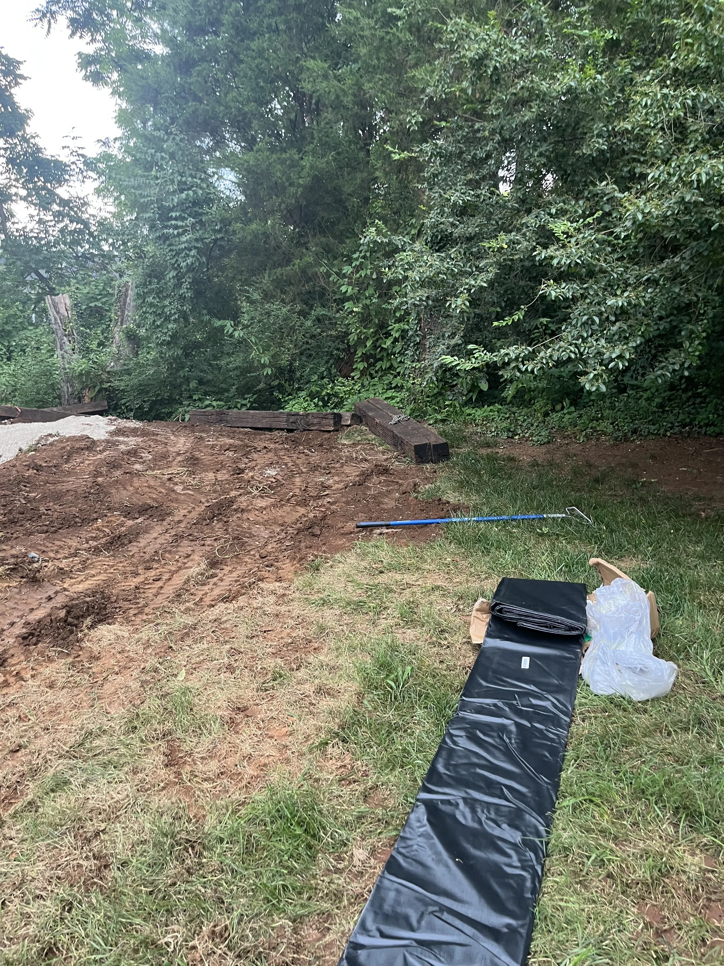 A cleared outdoor area with dirt and grass, some wooden planks, and a pile of black plastic sheeting, with trees in the background.
