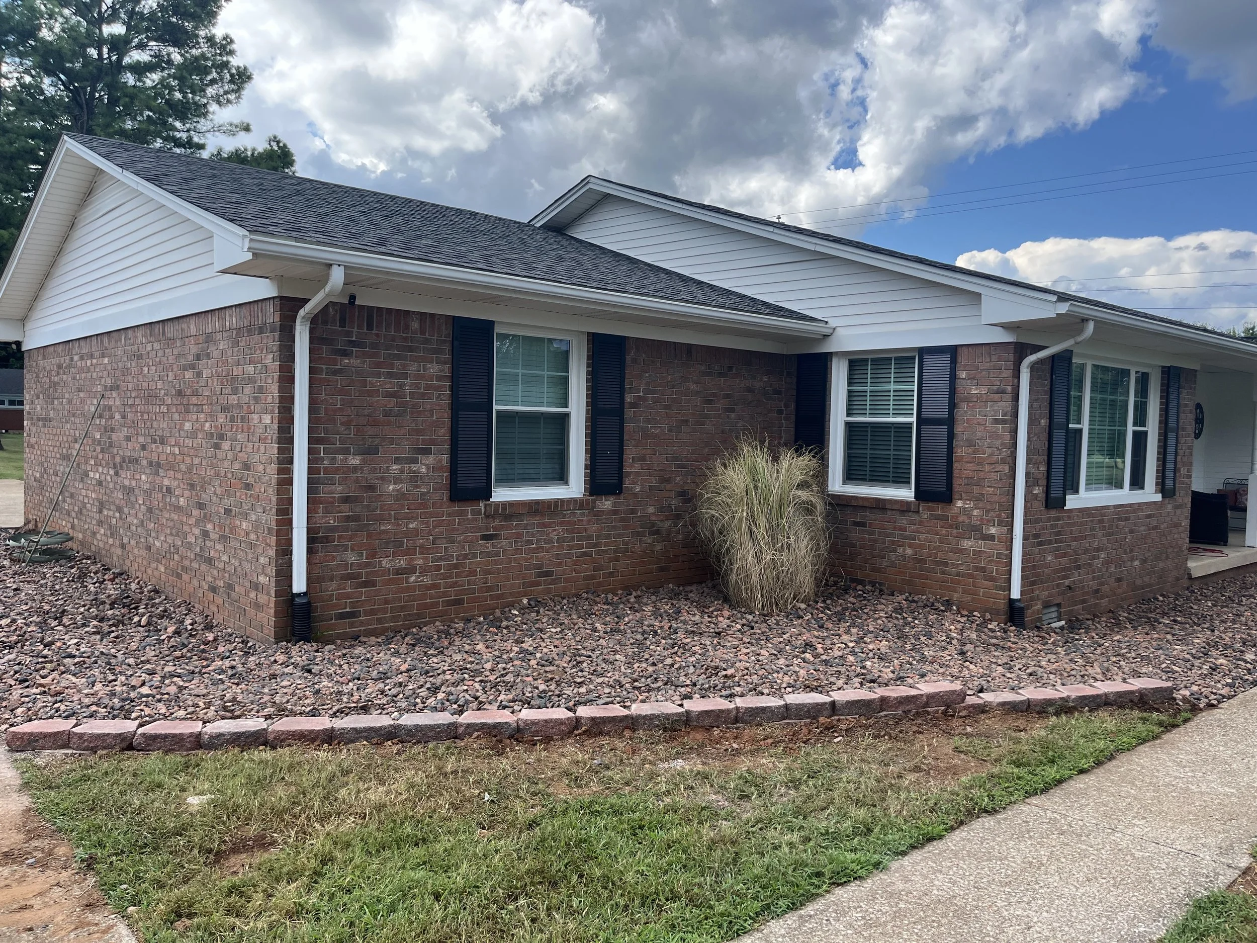 A single-story brick house with white siding, black shutters, and a gray shingled roof. There are three windows visible, and a small bush in front is surrounded by rocks. A concrete sidewalk runs along the front of the house.