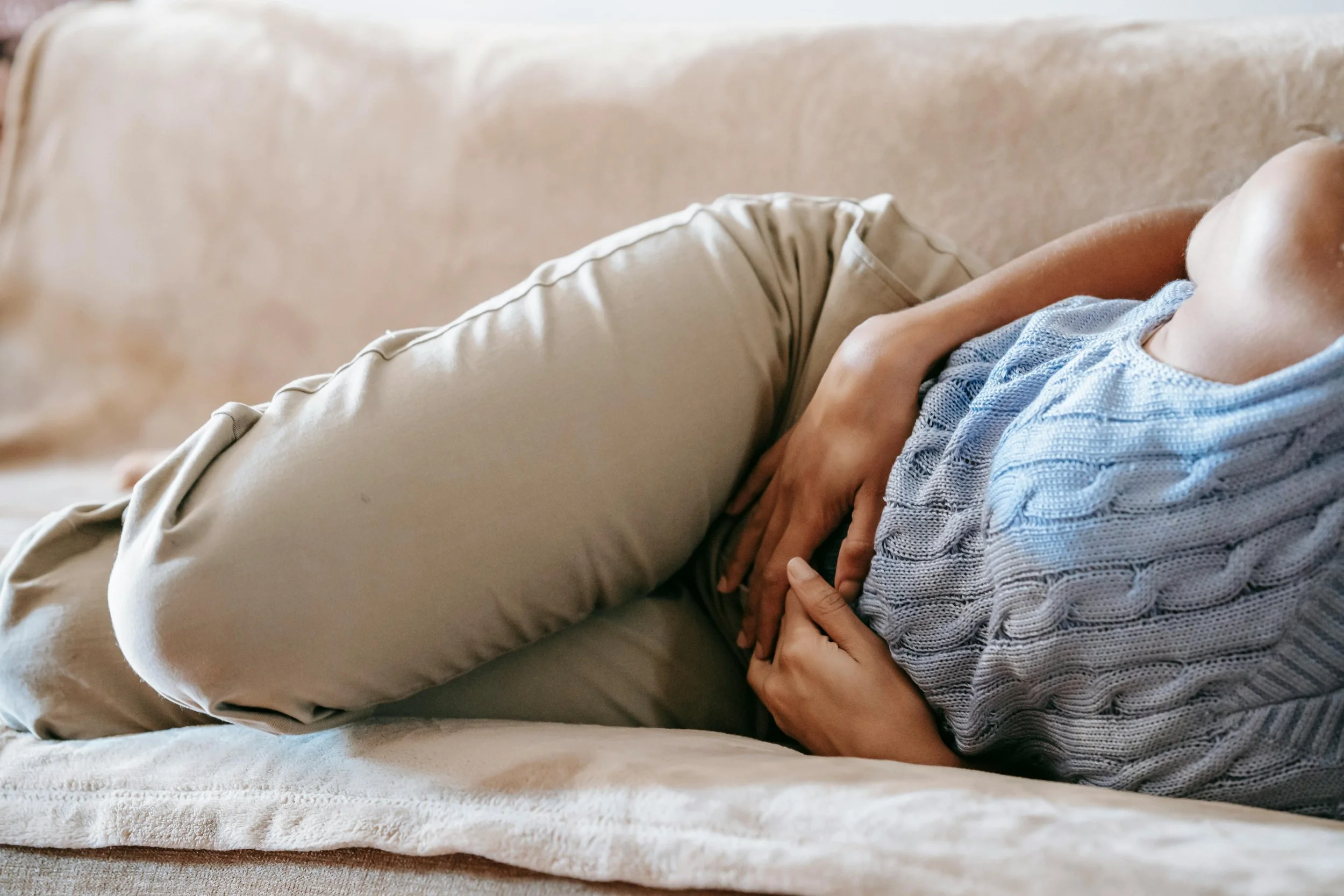 Person lying on a sofa with hands on stomach, wearing a blue knitted sweater and beige pants, in a relaxed posture.