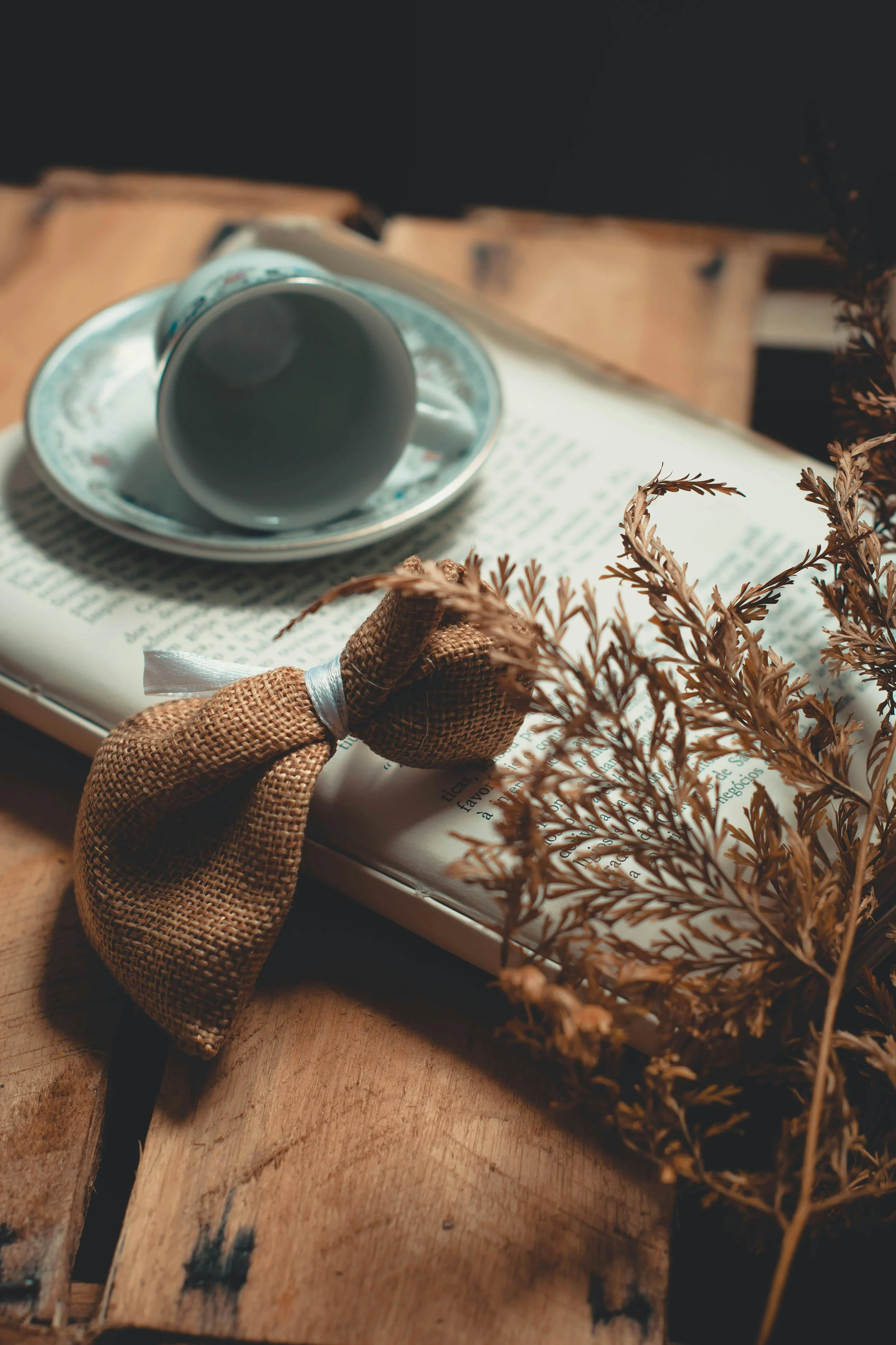 An open book with a teacup and saucer placed on it, a burlap bow, and dried plants on a wooden surface.