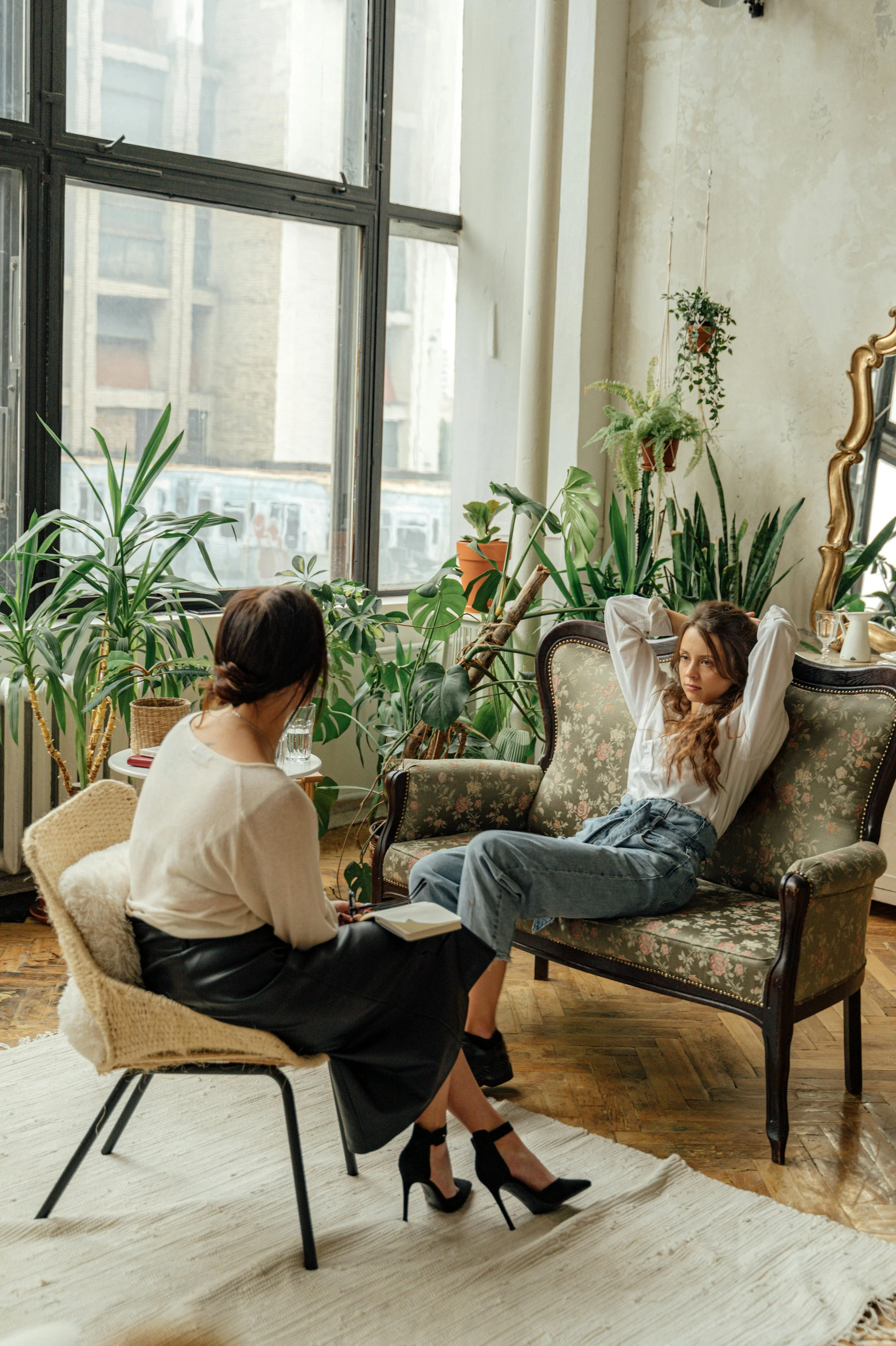 Two women having a conversation in a cozy, plant-filled room with large windows and vintage furniture.