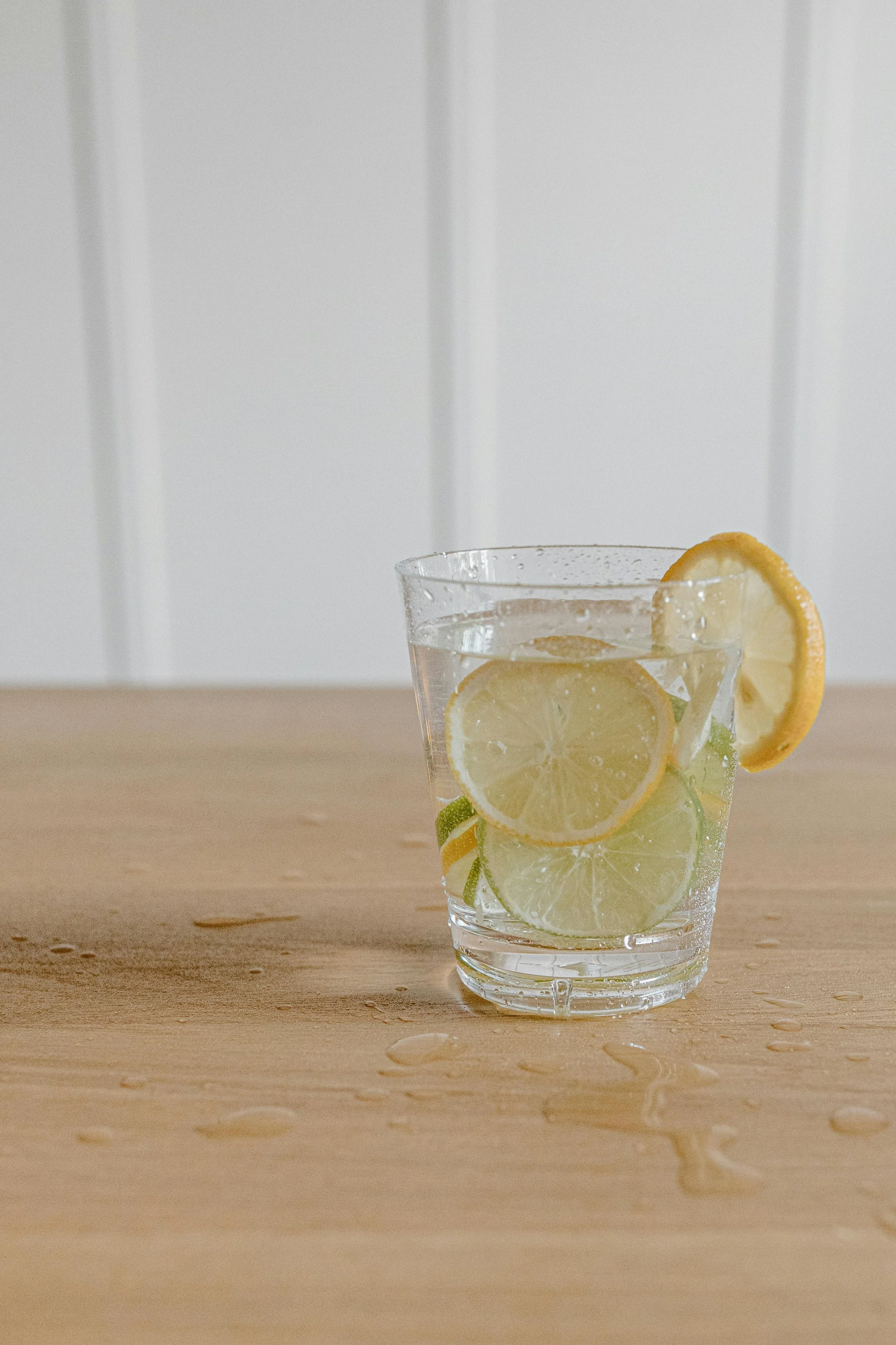 A clear glass of water with slices of lemon and lime, placed on a wooden surface with water droplets around it. A lemon slice decorates the rim of the glass. The background features a white paneled wall.