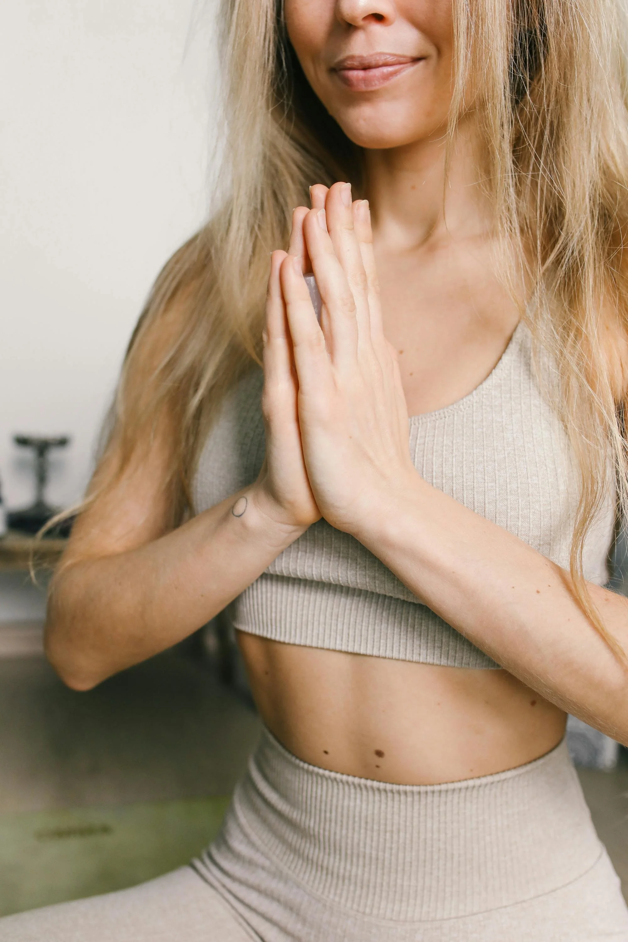 Close-up of a woman with long blonde hair practicing yoga indoors, holding her hands together in a prayer position near her chest, wearing a beige sports bra and matching high-waisted leggings.